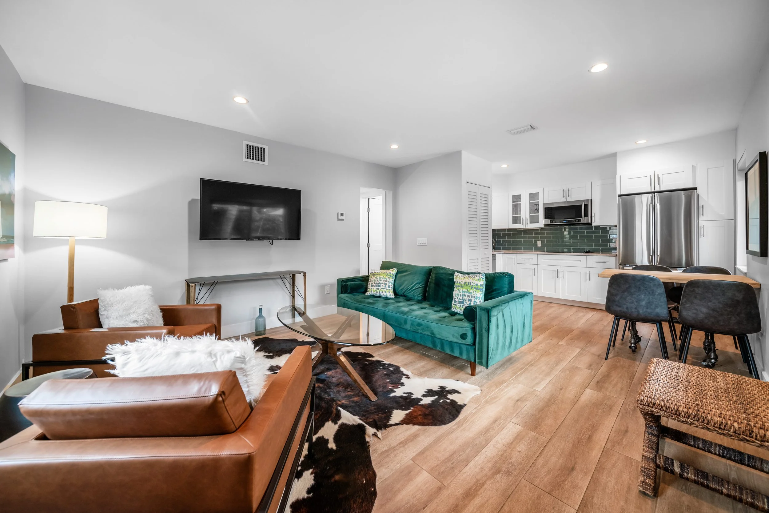 Open-concept living room and kitchen with teal sofa, brown leather accent chairs, cowhide rug, wall-mounted TV, kitchen with white cabinets, green subway tile backsplash, stainless steel appliances, and wooden dining table with black chairs.