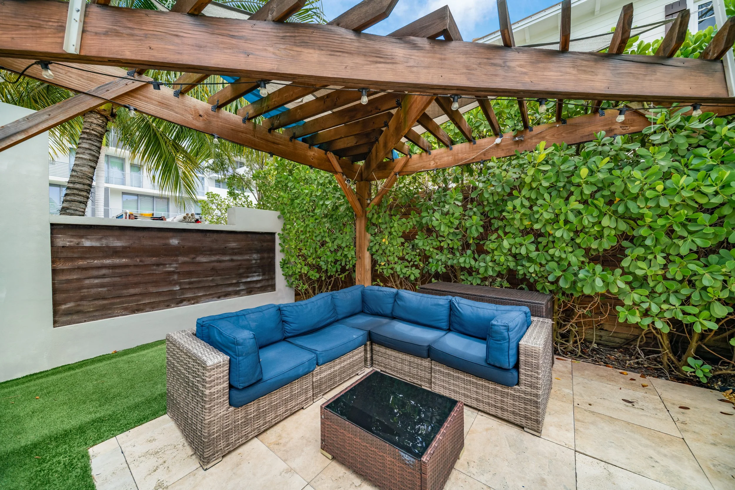 Outdoor patio with a wicker sectional sofa with blue cushions, a small black glass-top table, surrounded by green plants and trees, under a wooden pergola with string lights.