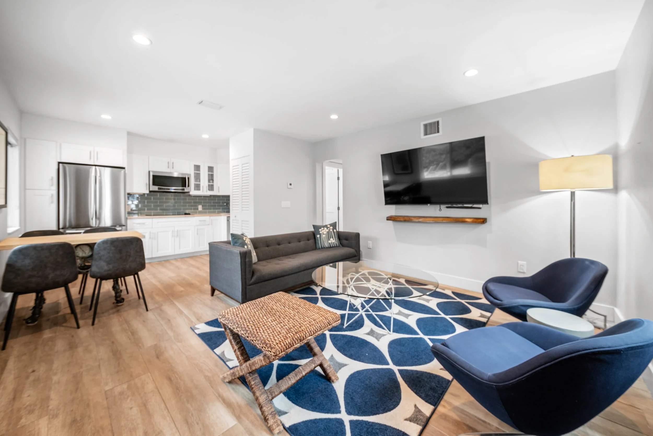 Living room with a gray sofa, dark blue chairs, a colorful rug, a flat-screen TV on the wall, floor lamp, and an open kitchen with white cabinets and stainless steel appliances.