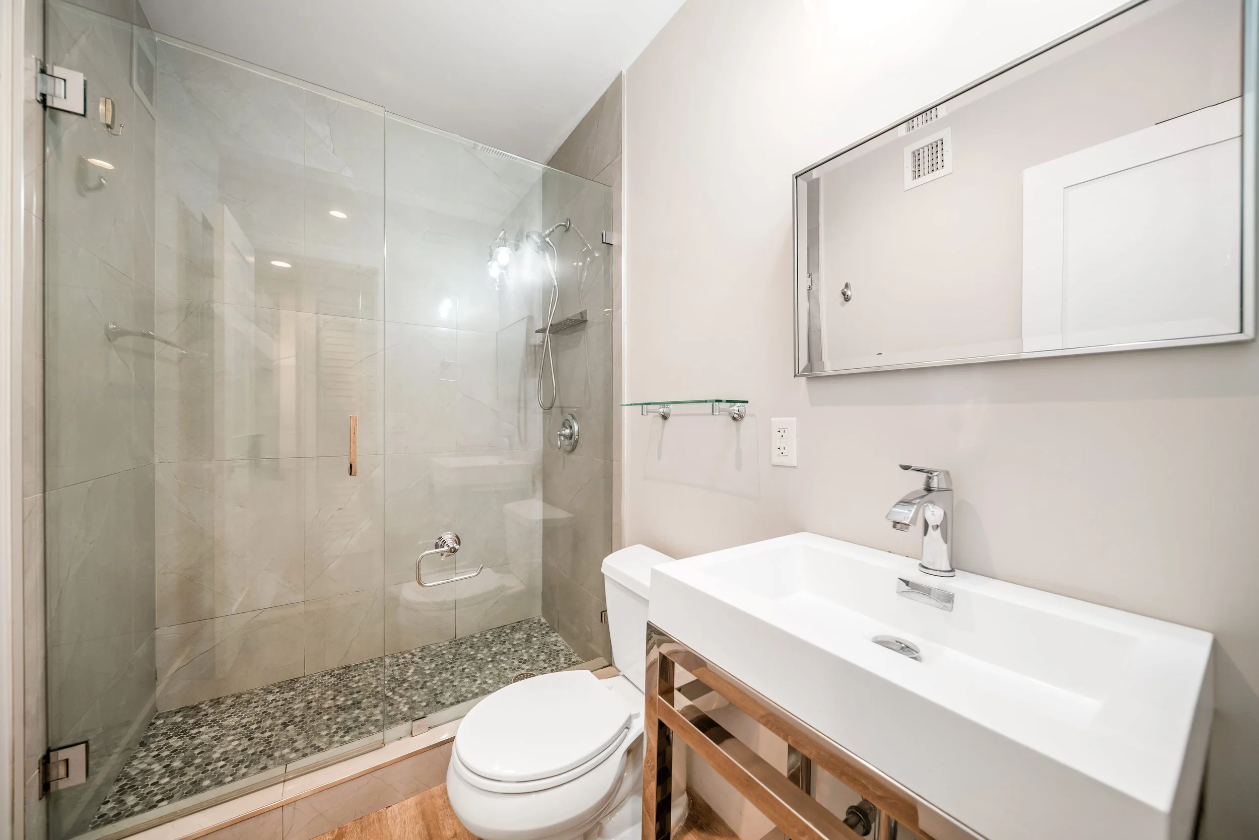 Bathroom with a glass-enclosed shower, a toilet, a white sink with a wooden stand, a rectangular mirror, and a glass shelf, all in a modern minimalist style.
