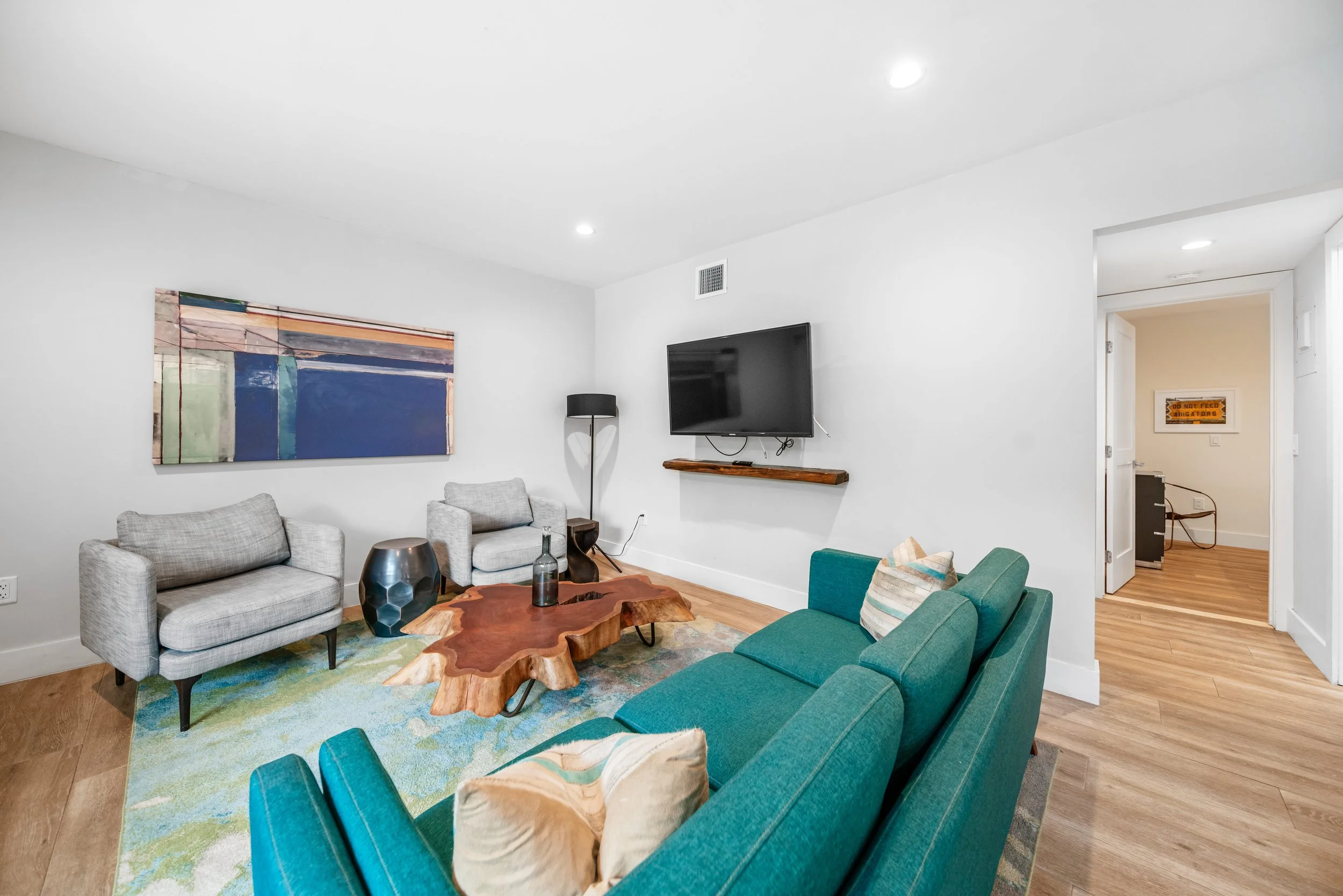 Living room with gray and teal furniture, a wooden coffee table, wall-mounted TV, abstract wall art, and hardwood flooring.