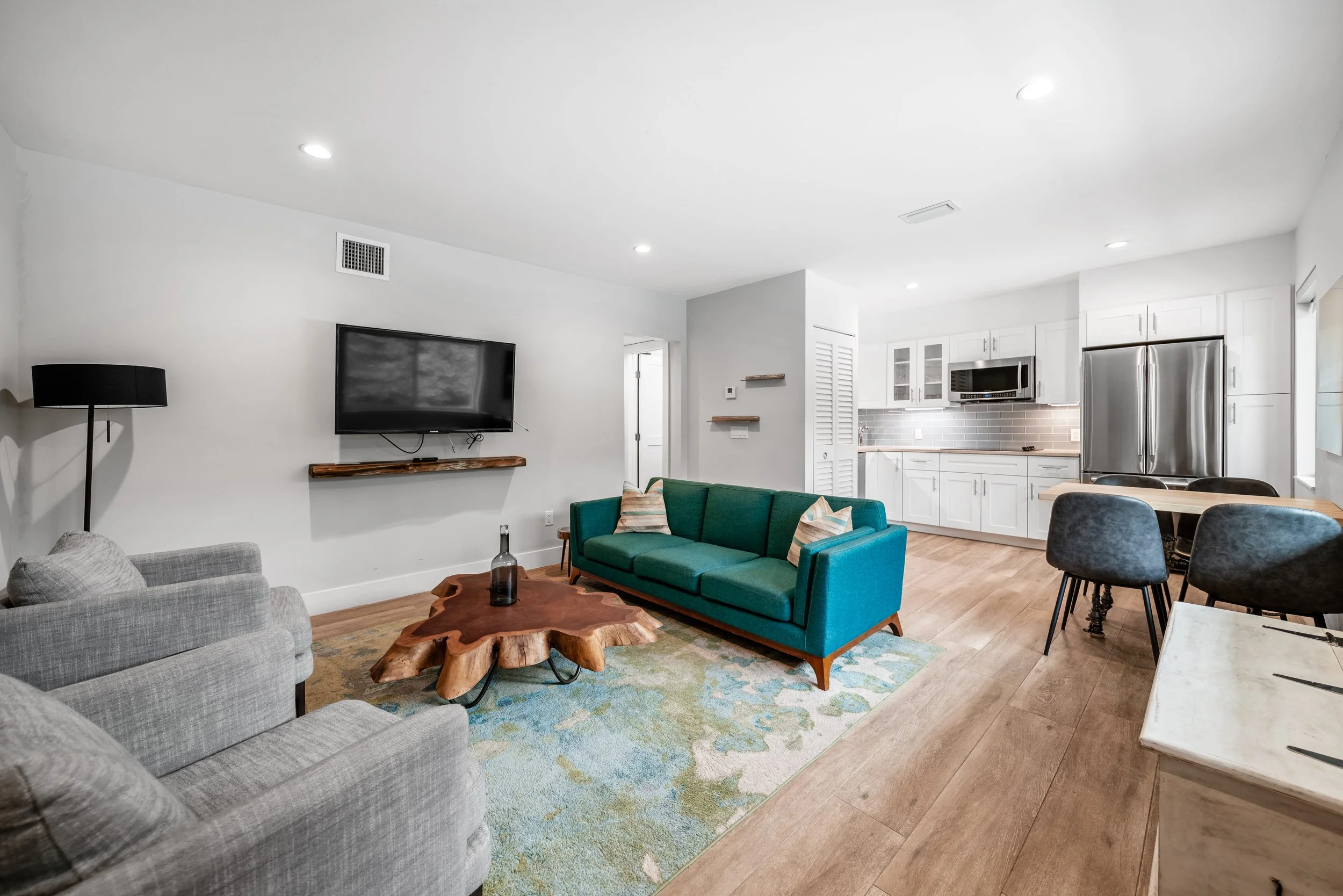 Living room with gray and teal sofas, a wooden coffee table, a wall-mounted TV, a floor lamp, and an open kitchen with white cabinets, a stainless steel refrigerator, and grey backsplash.
