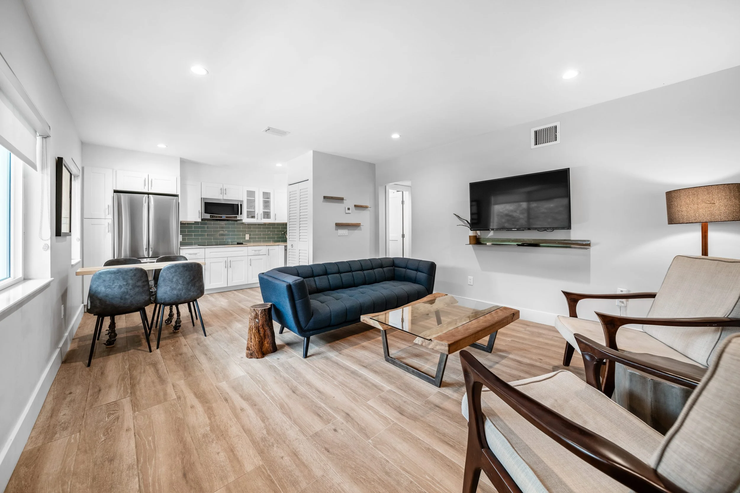 Living room with a blue couch, beige armchairs, a wooden coffee table, a wall-mounted TV, and a kitchen with white cabinets and stainless steel refrigerator in the background.