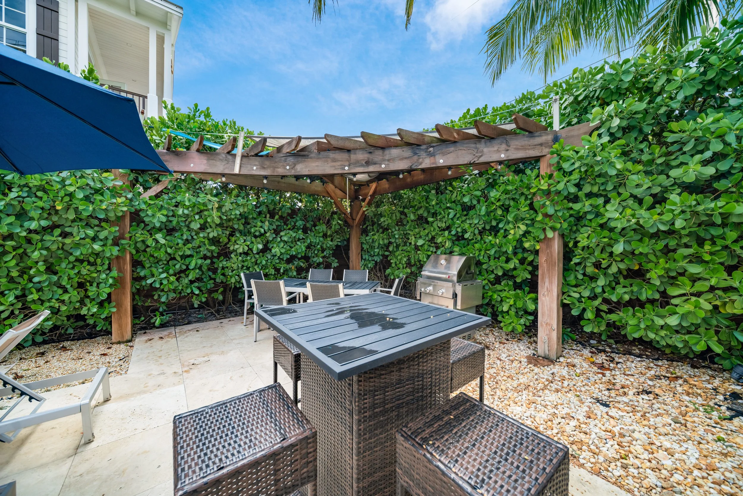 Outdoor patio with patio chairs, a grill, a blue umbrella, and lush green bushes under a clear sky.
