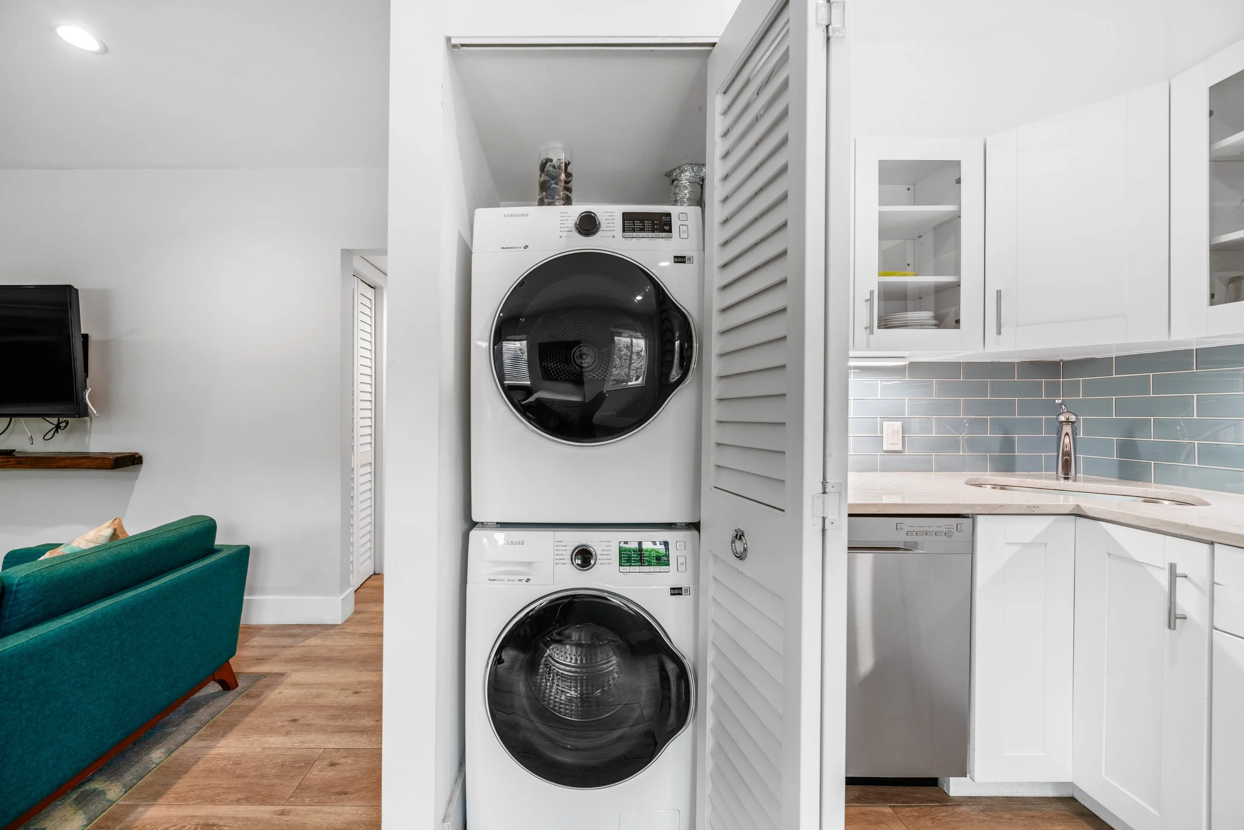 Stacked washer and dryer in a laundry closet next to a kitchen with white cabinets and a blue subway tile backsplash.