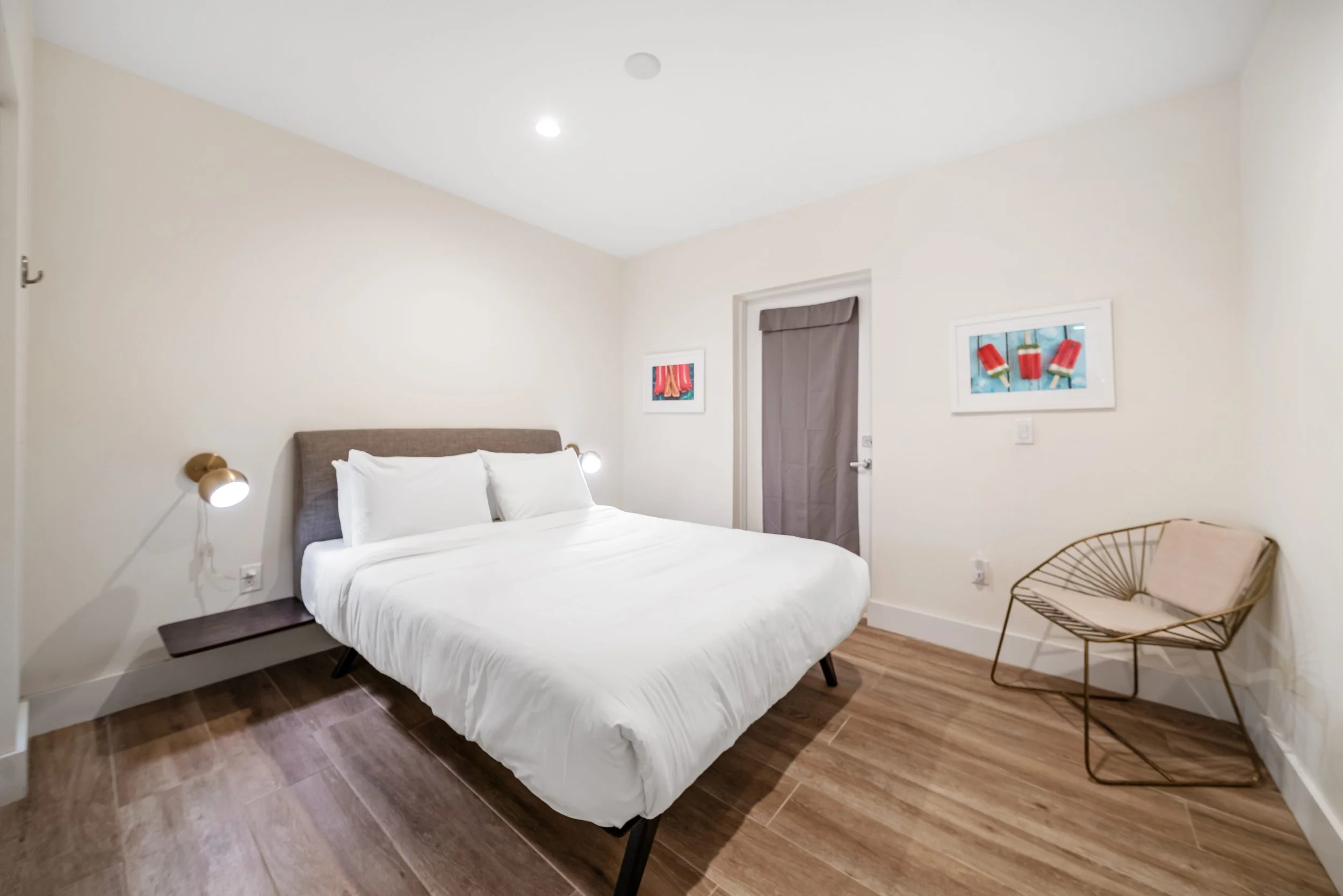A modern bedroom with a bed, white bedding, two wall-mounted lamps, two framed colorful art pieces, a metallic chair with a cushion, and a door with a curtain.