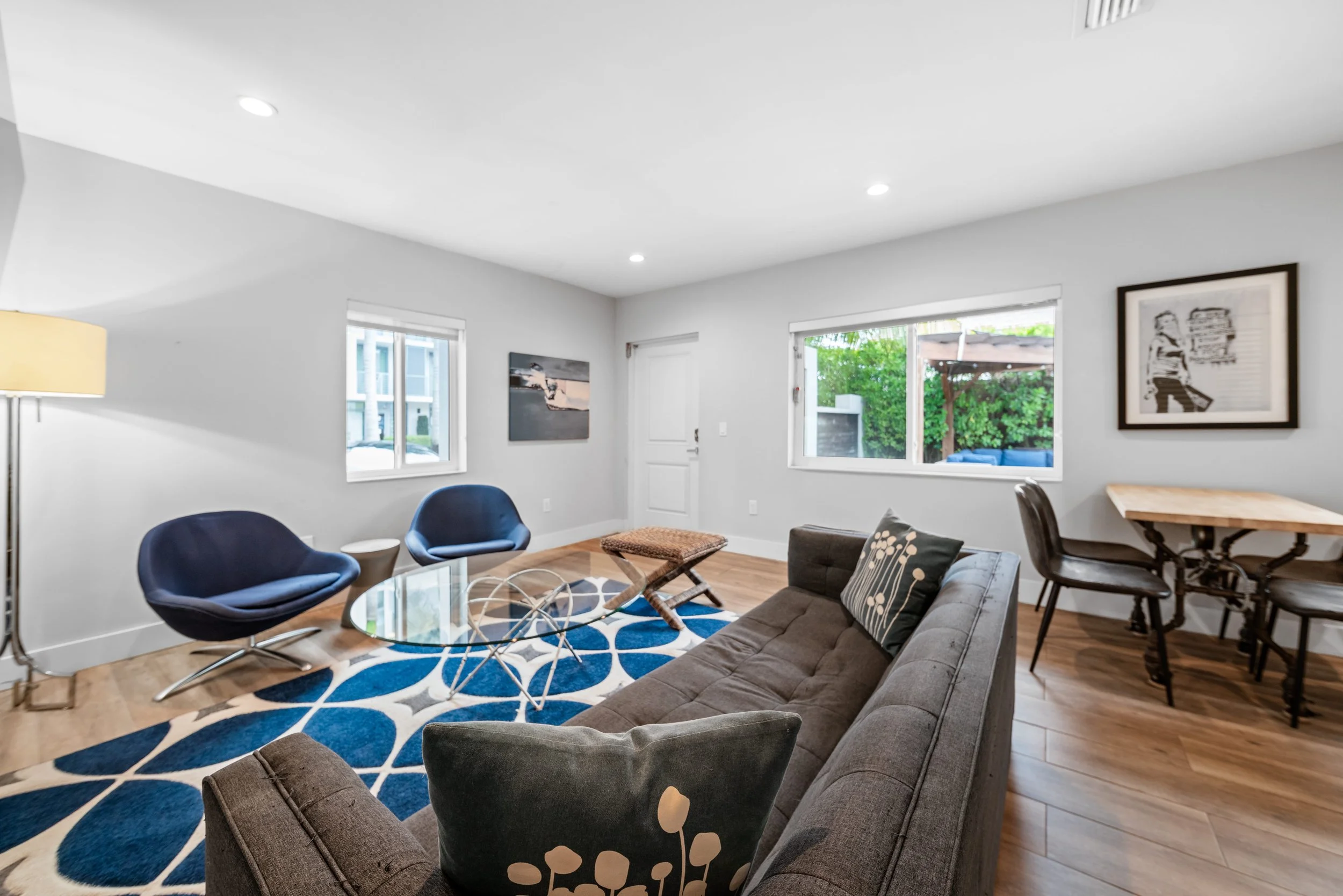 Living room with hardwood floors, white walls, a brown sofa with patterned pillows, two blue chairs around a glass coffee table, a rug with blue and white pattern, a small dining area with a wooden table and chairs, outdoor view through windows, fram
