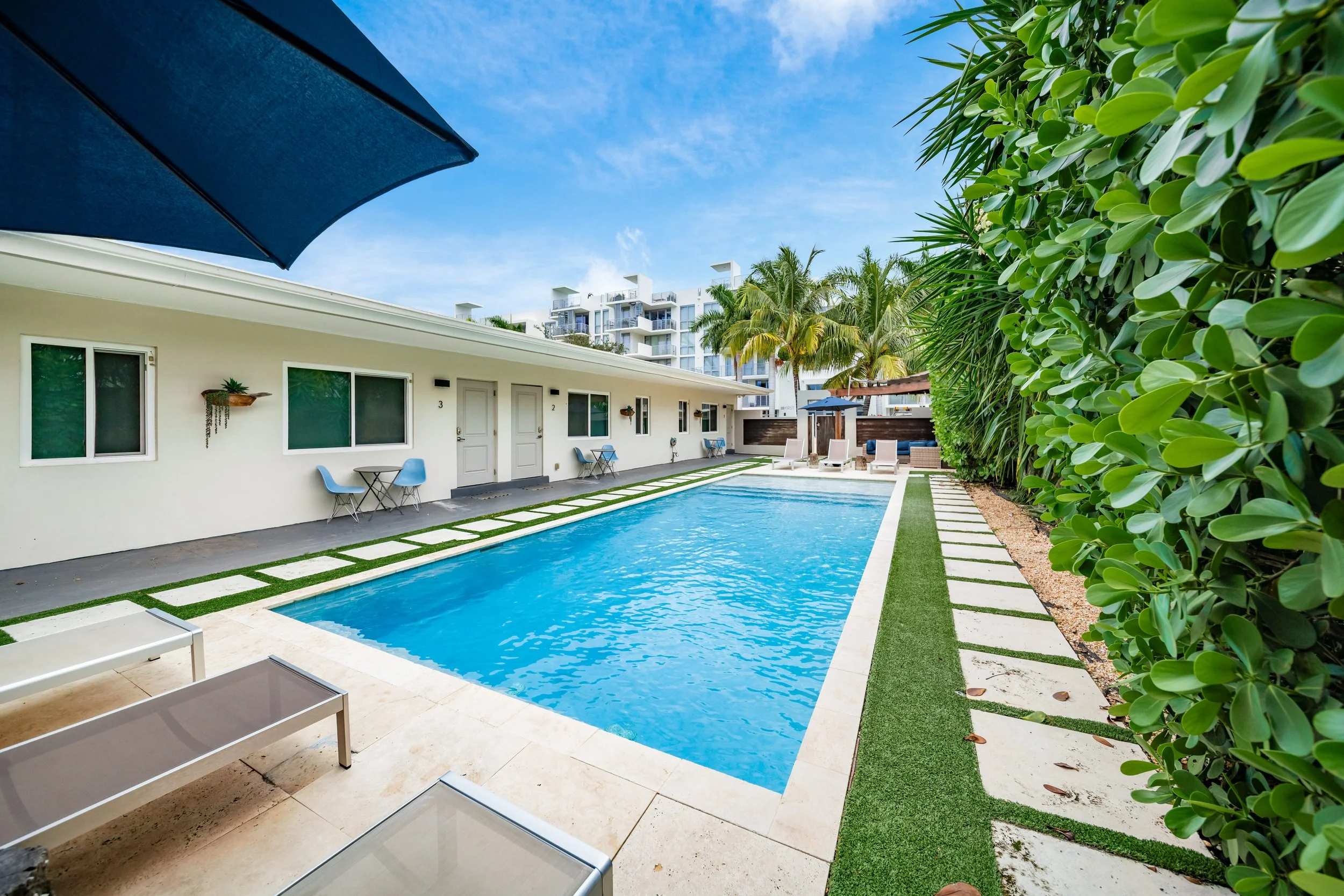 A backyard swimming pool surrounded by a white building with multiple windows, lounge chairs, palm trees, and lush greenery under a blue sky.