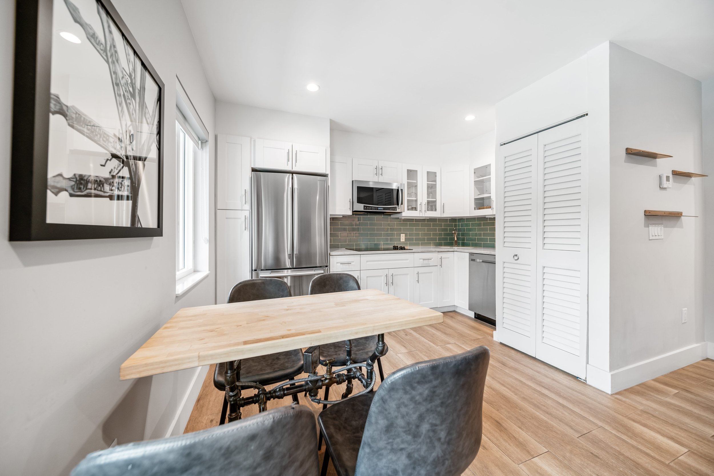 Modern kitchen with white cabinets, stainless steel refrigerator, green subway tile backsplash, microwave, and dining table with chairs.