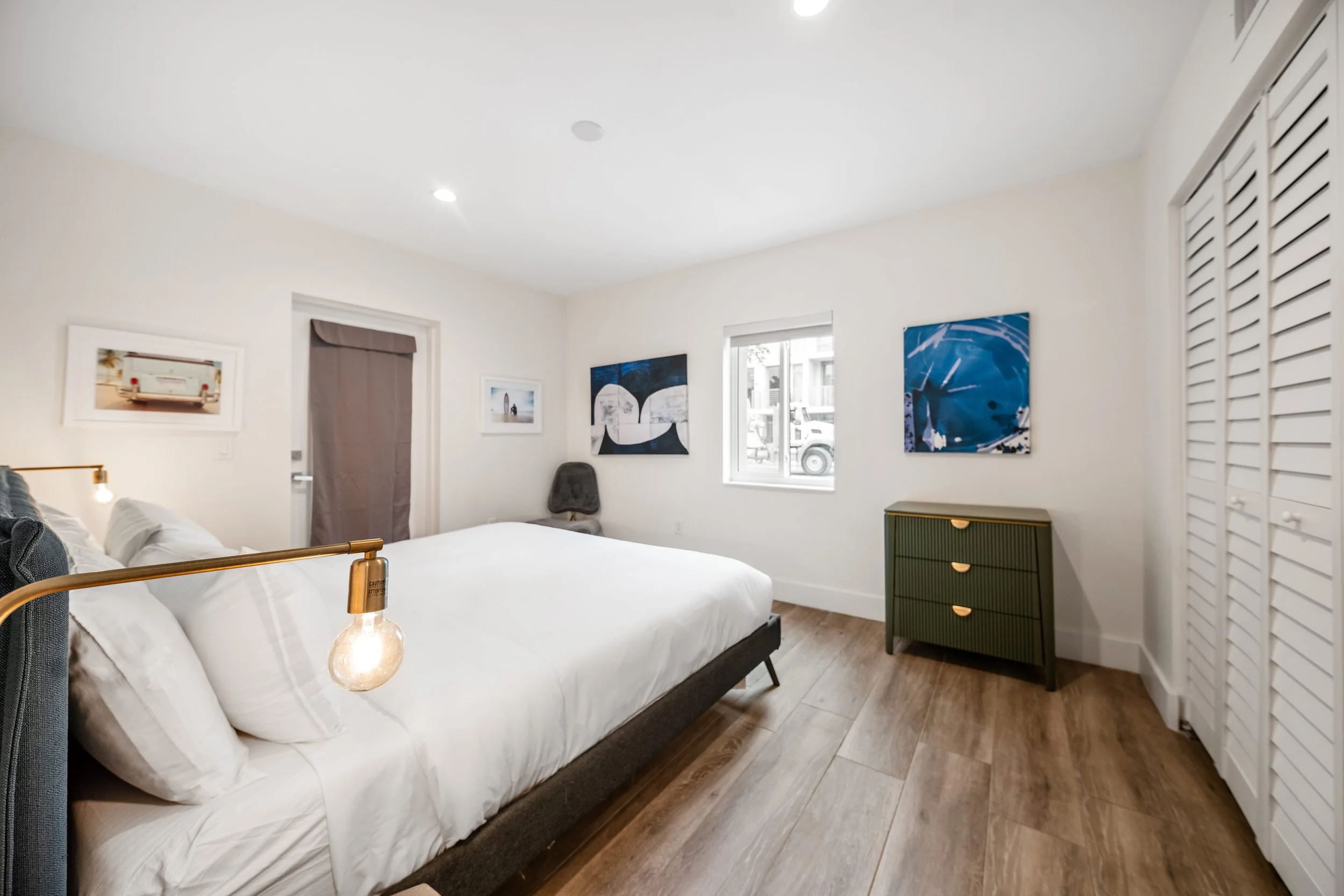 A modern bedroom with white walls, wooden flooring, a bed with white bedding and pillows, and a brass reading lamp attached to the bed frame. There is a green dresser with three drawers, abstract blue artwork on the walls, a window, and a gray chair 