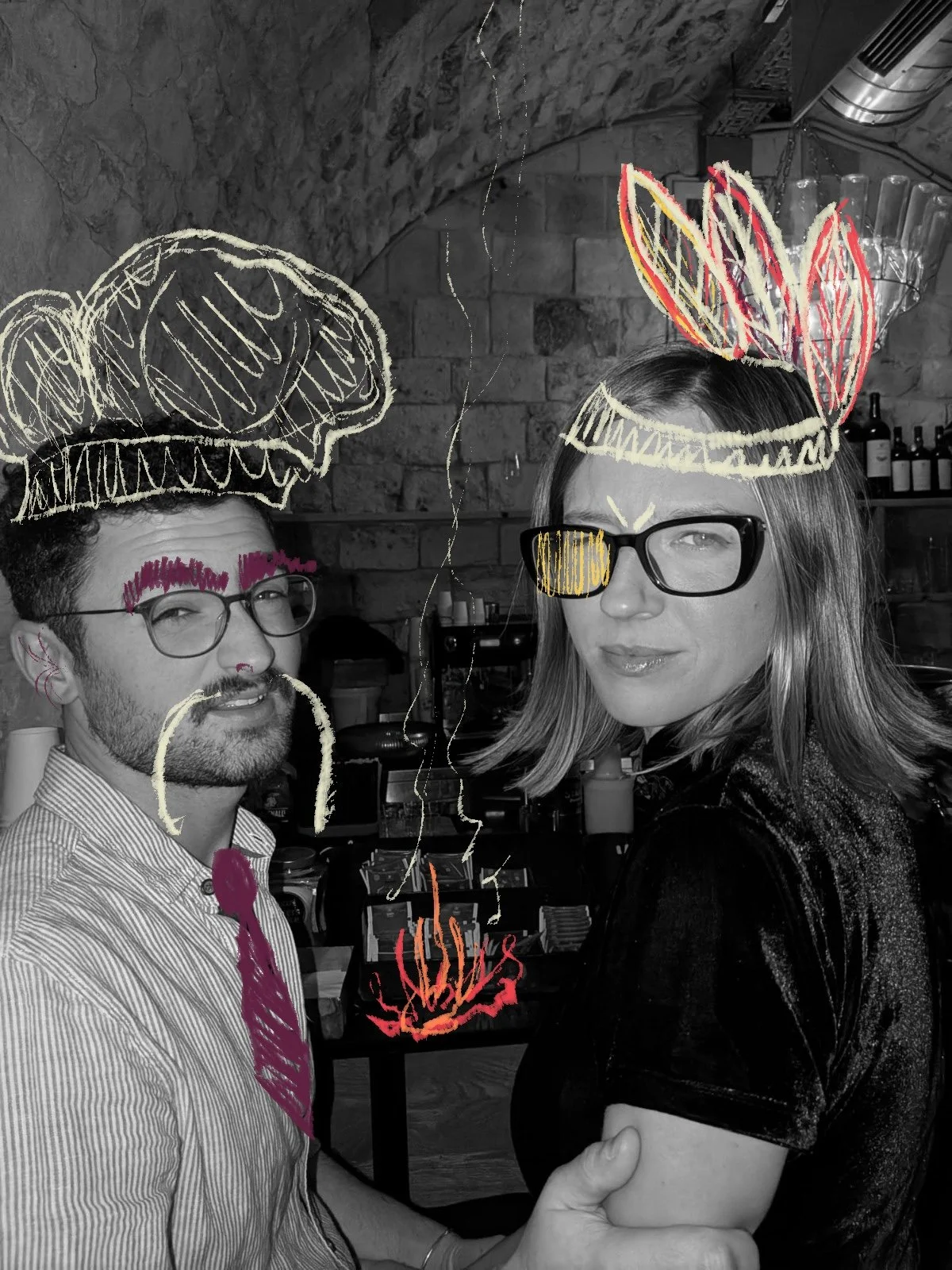 Two people are in a black and white photograph with drawn colorful doodles over their faces and heads. The person on the left is a man with glasses, a beard, and a striped shirt, with pink doodles on his glasses, eyebrows, and a mustache. The person on the right is a woman with glasses and shoulder-length hair, with yellow, red, and pink doodles on her head and face. The background shows a brick wall and shelves with bottles and kitchen items.