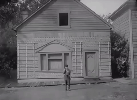 A person standing in front of a small, old wooden house with a front door and a large window. The house appears to be under construction or renovation, with exposed framing and no paint.