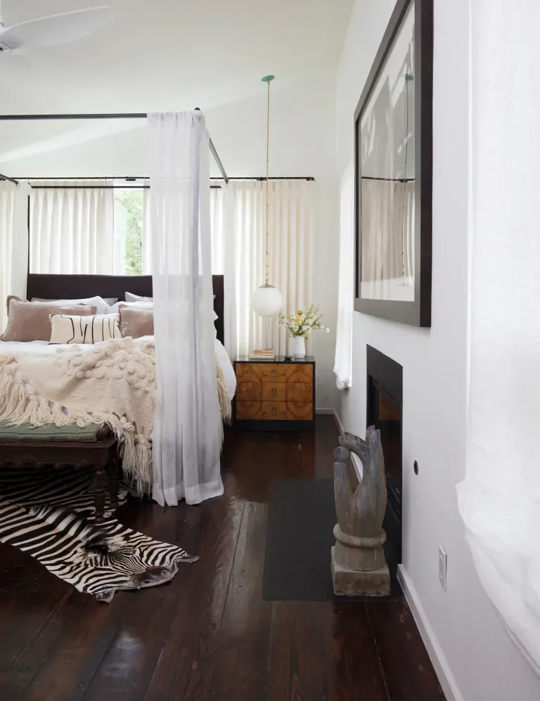 Soft textiles, warm wood flooring, and filtered light establish the bedroom as a private retreat. The canopy frame and understated fireplace anchor the room with subtle drama, while every detail supports comfort and calm.