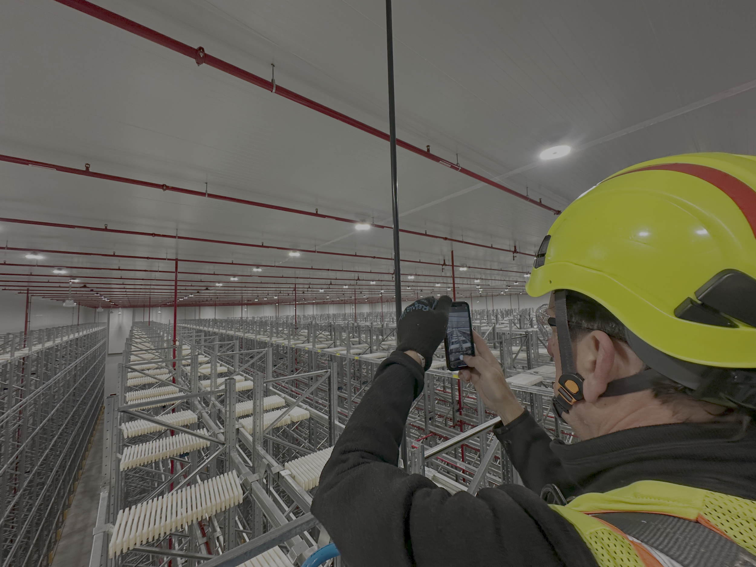 Una persona con casco amarillo y guantes toma una foto en un almacén de estanterías metálicas con productos industriales.