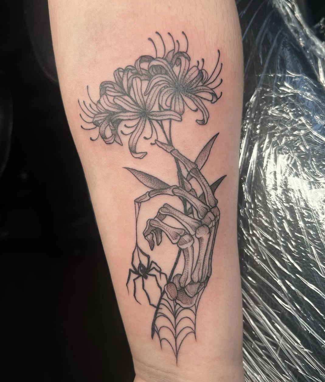 A tattoo of a skeletal hand holding a flower with spider and web details in a black outline style.