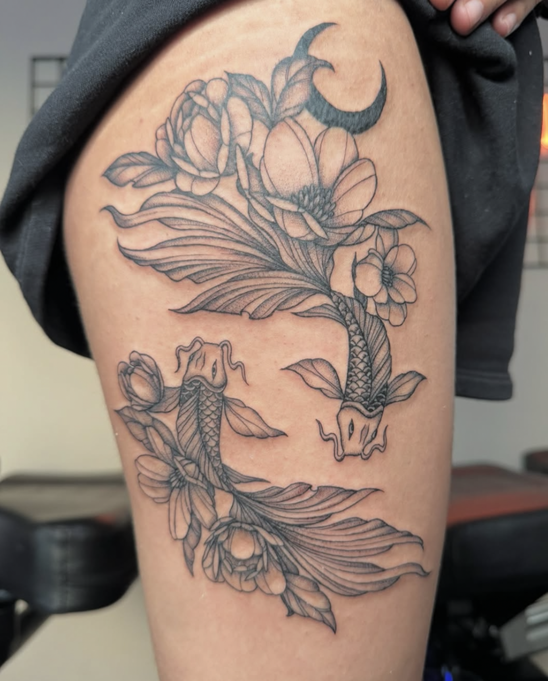 Black and gray tattoo of peonies, leaves, a crescent moon, and two koi fish on person's upper arm.