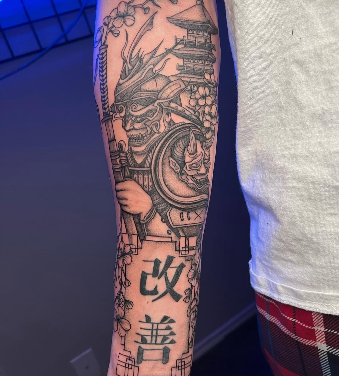 Tattoo of a Japanese warrior holding a spear, with a pagoda and cherry blossoms, along with Chinese characters on the forearm.