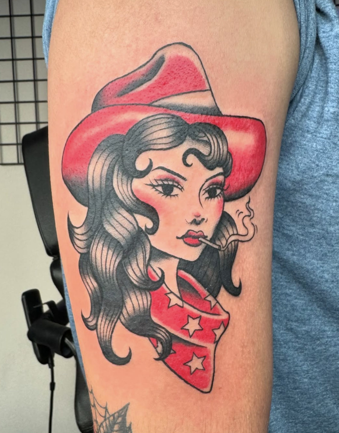 Tattoo of a woman with dark wavy hair wearing a red wide-brimmed hat, with makeup and a cigarette, on upper arm.