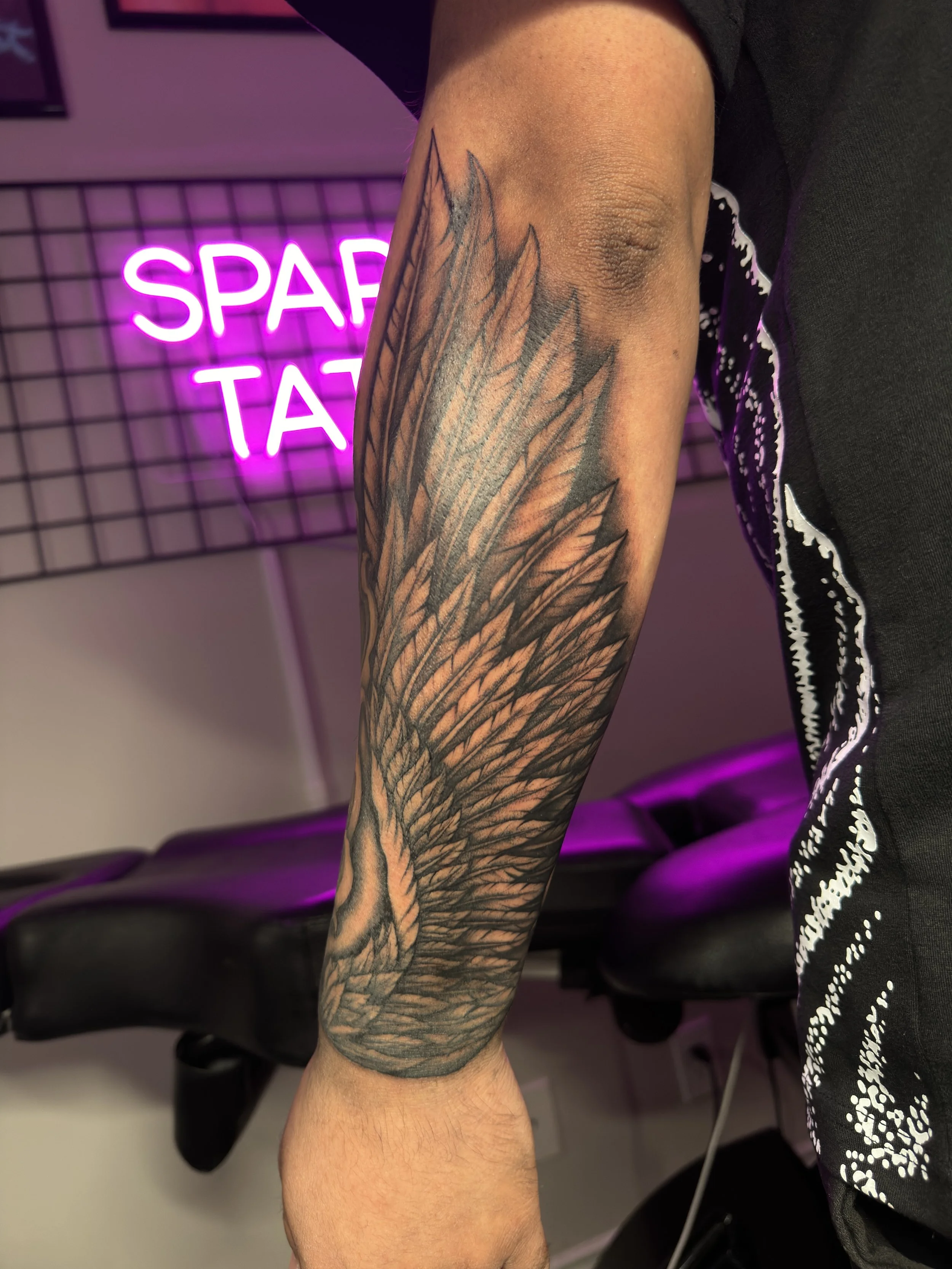 A person's arm with a detailed black and gray tattoo of angel wings on the forearm.