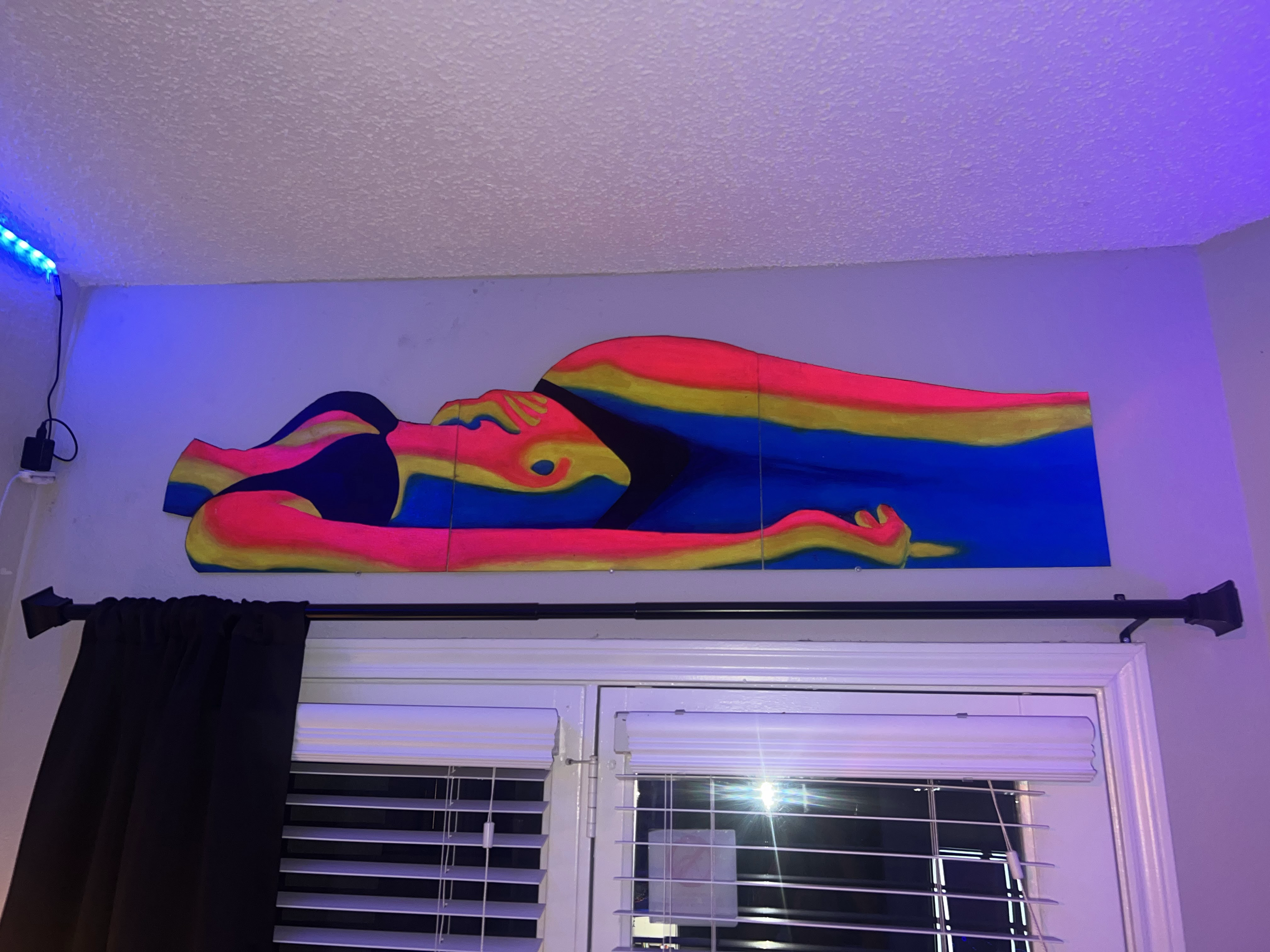 Colorful abstract painting of a reclining woman hanging on a wall above a window with black curtains.