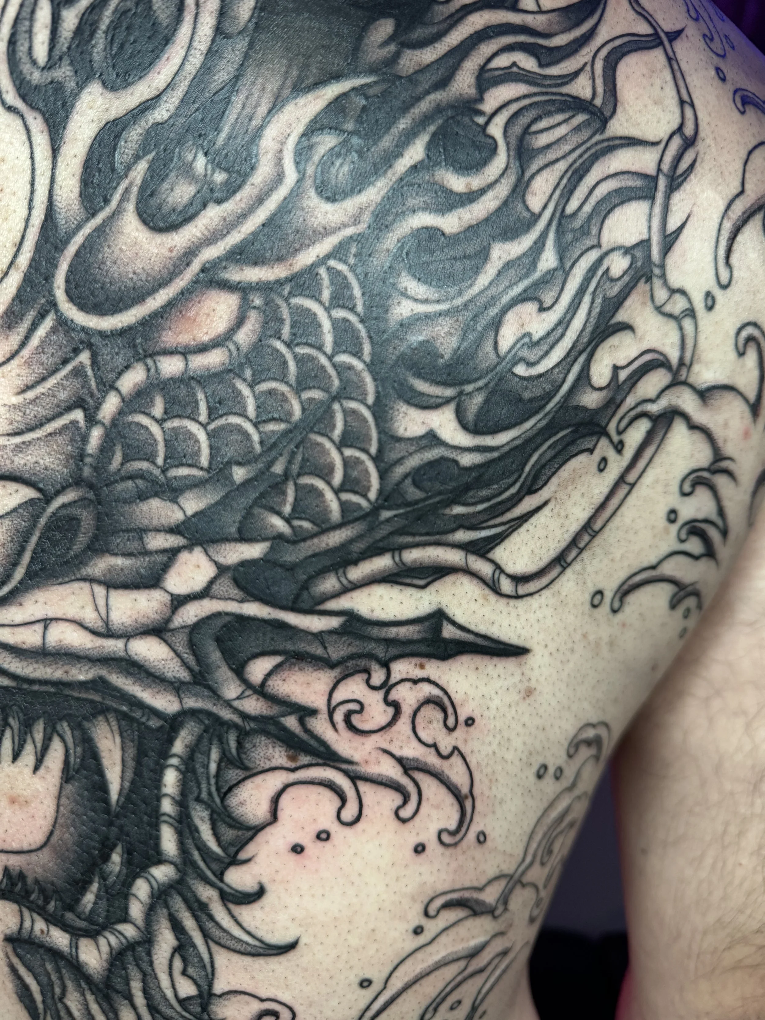 Close-up of a black ink tattoo depicting a tree, clouds, and waves on a person's shoulder and upper arm.