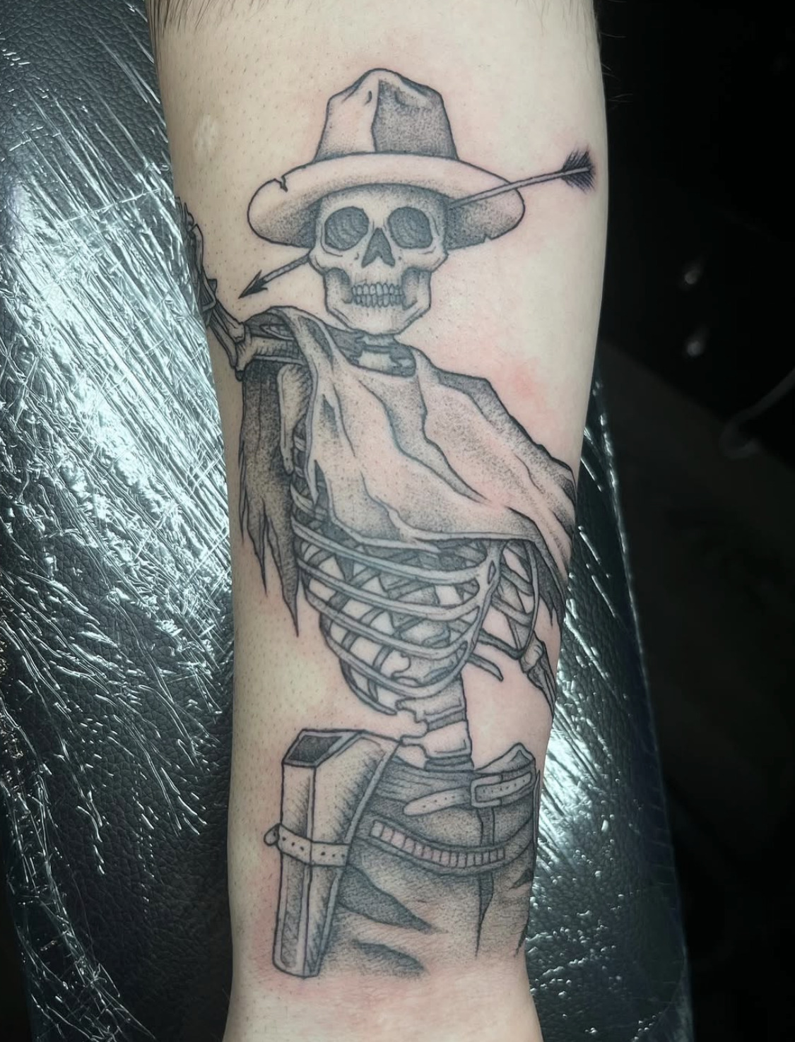 Tattoo of a skeleton wearing a hat with an arrow through its skull, dressed in a shirt, with a ribcage and firearm on its side.