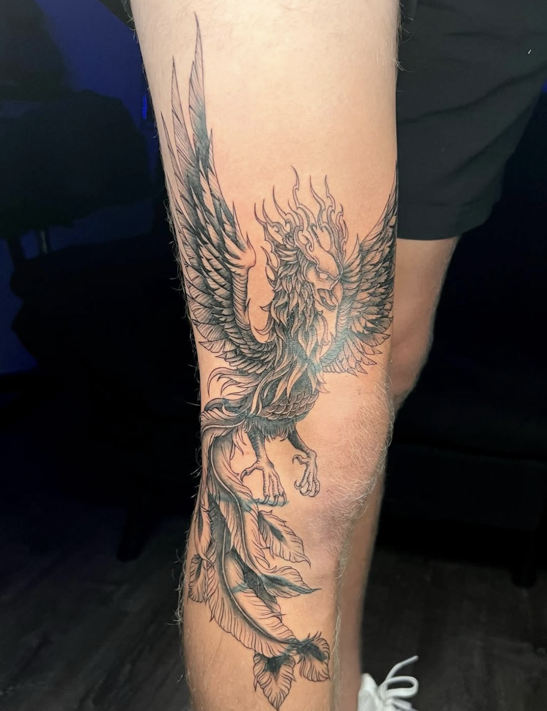 Black and gray detailed tattoo of a phoenix with wings spread, perched on a leafy branch.