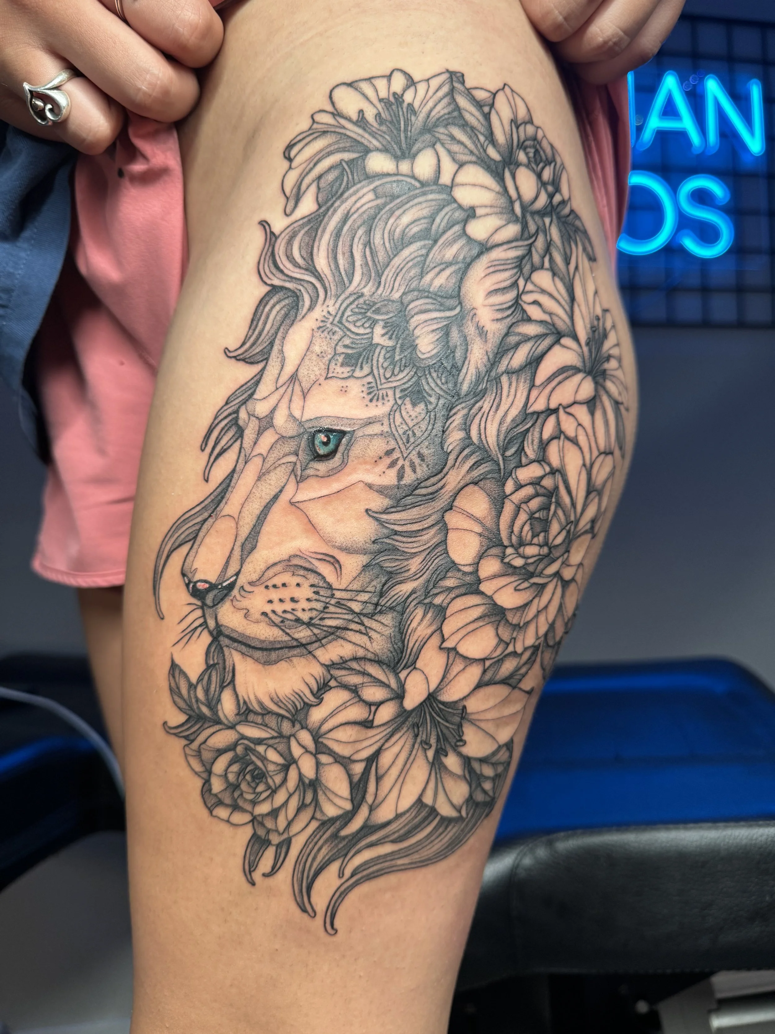 A tattoo of a lion's face surrounded by detailed flowers on a person's thigh.