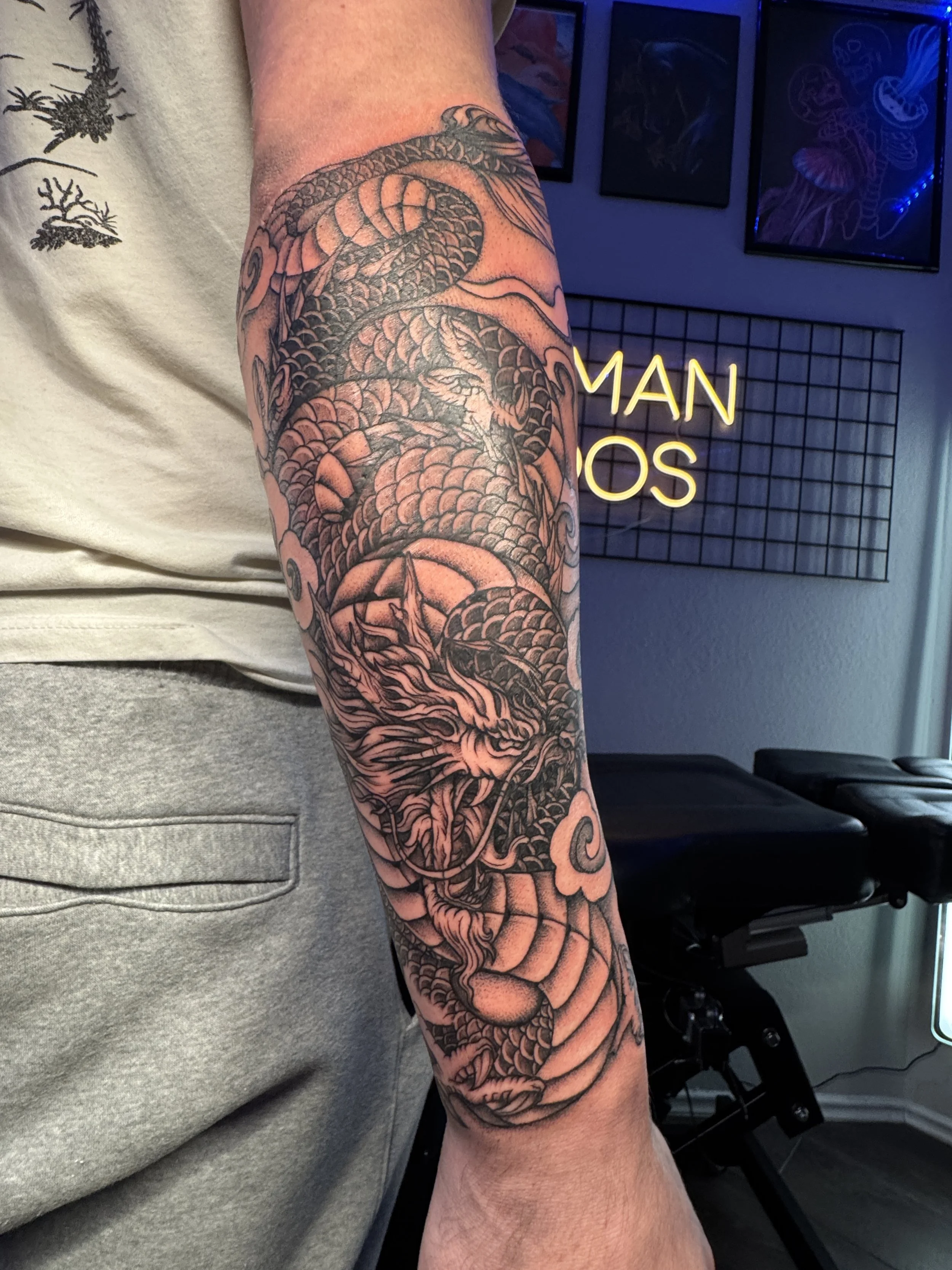 Black and gray tattoo of a dragon on a person's forearm with clouds and scales.