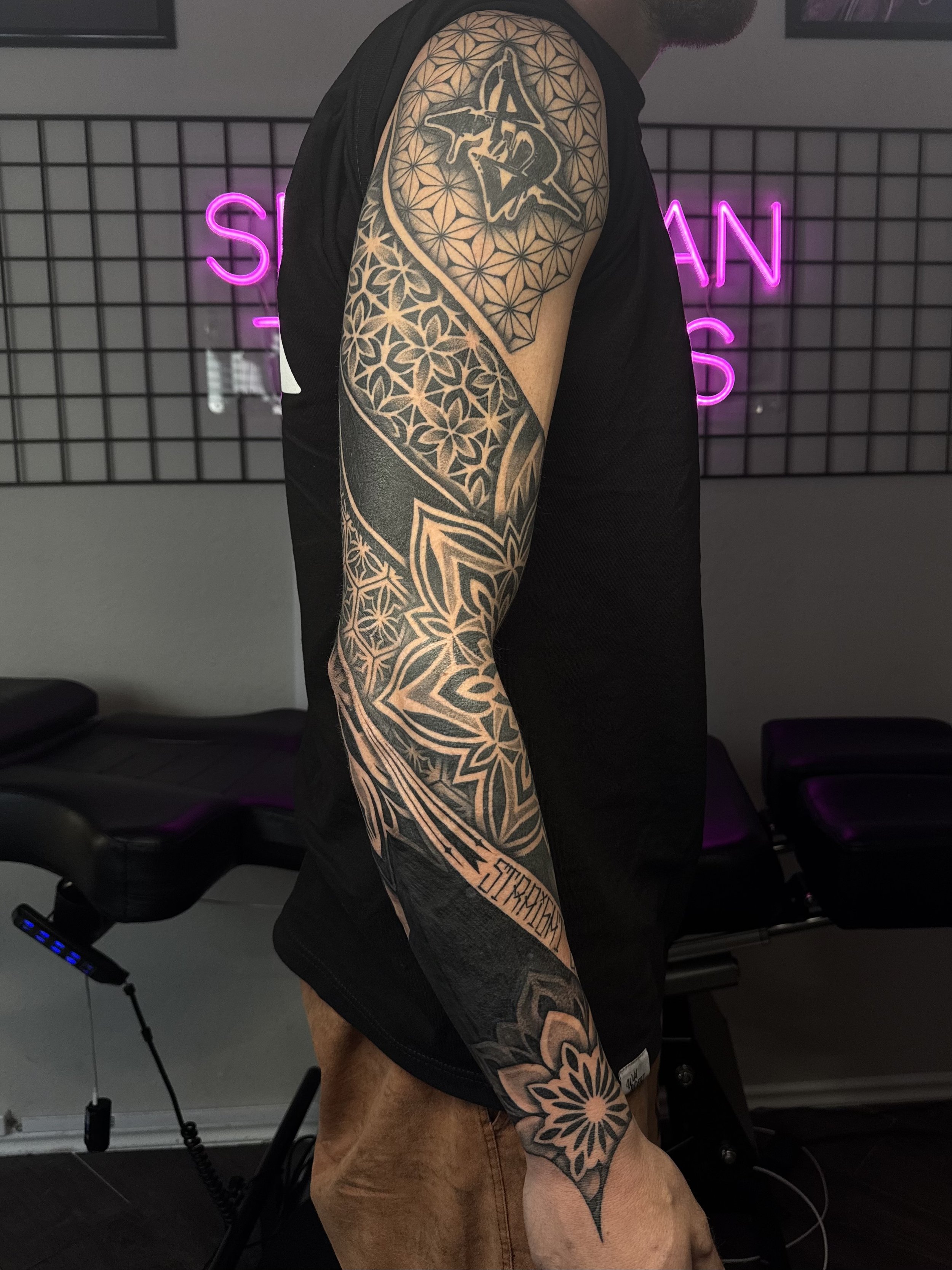 A person with a full sleeve tattoo on their arm, featuring geometric patterns, floral designs, and text, standing in a tattoo studio with neon pink and purple lighting.