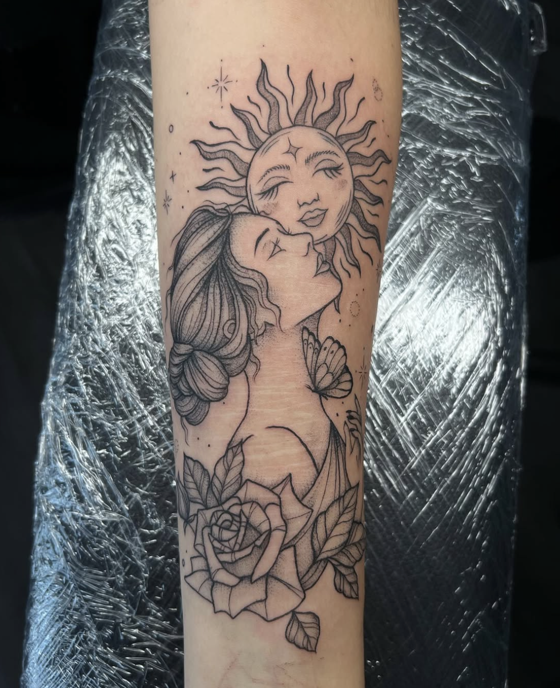 Tattoo of a sun with a smiling face and closed eyes, personified with long eyelashes, above a woman with long hair kissing the moon, surrounded by a butterfly, rose, and leaves.