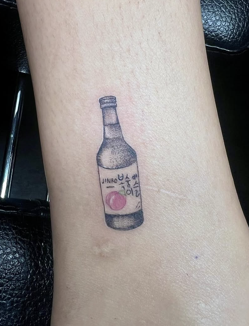 Tattoo of a small bottled beverage on a person's arm, with Korean writing on the label and a pink circular logo.