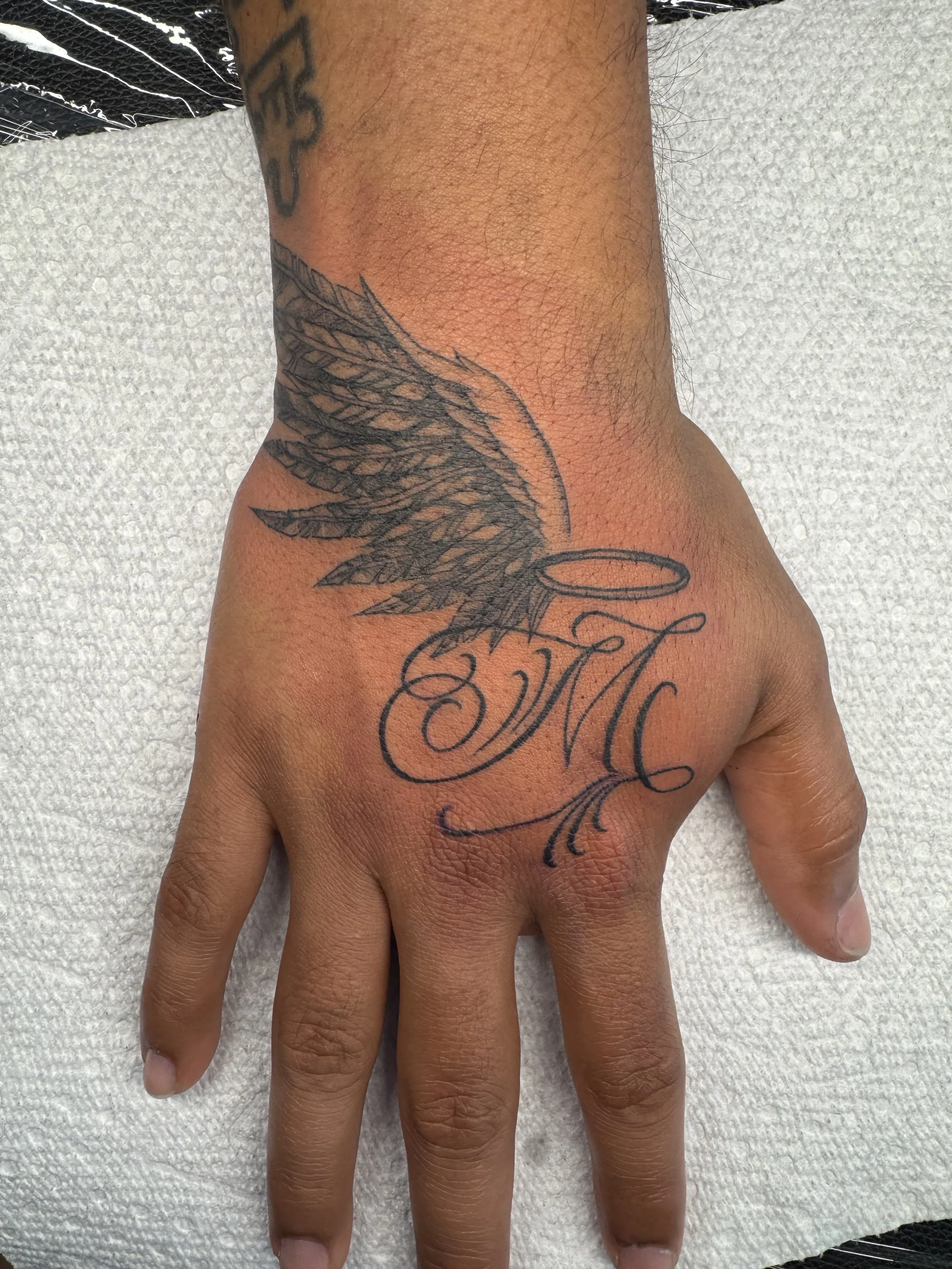 A person's hand with a tattoo of a wing and a halo above cursive initials 'AMC' on the top of the hand.