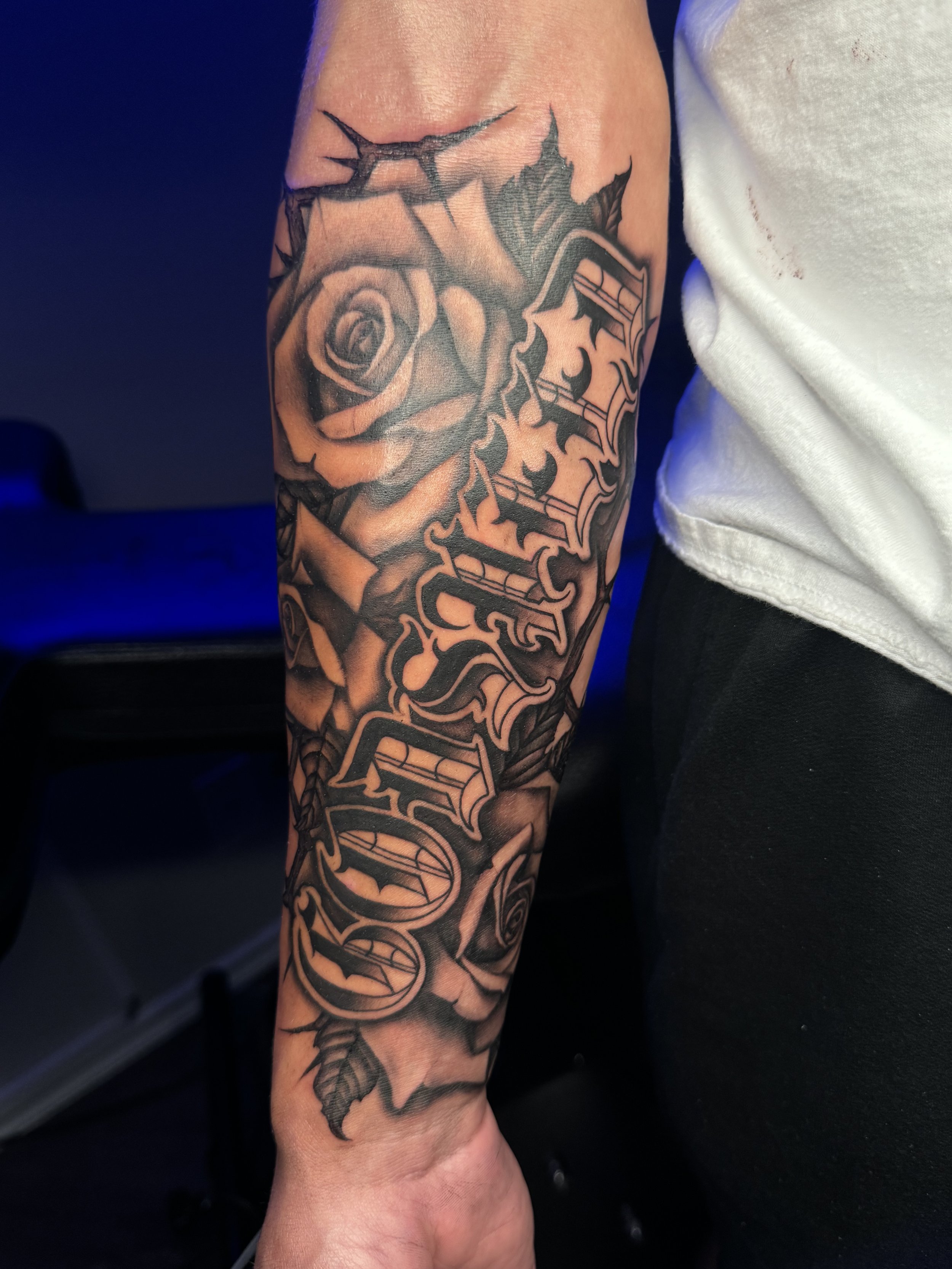 Tattoo of a large rose and gothic-style text on a person's forearm.