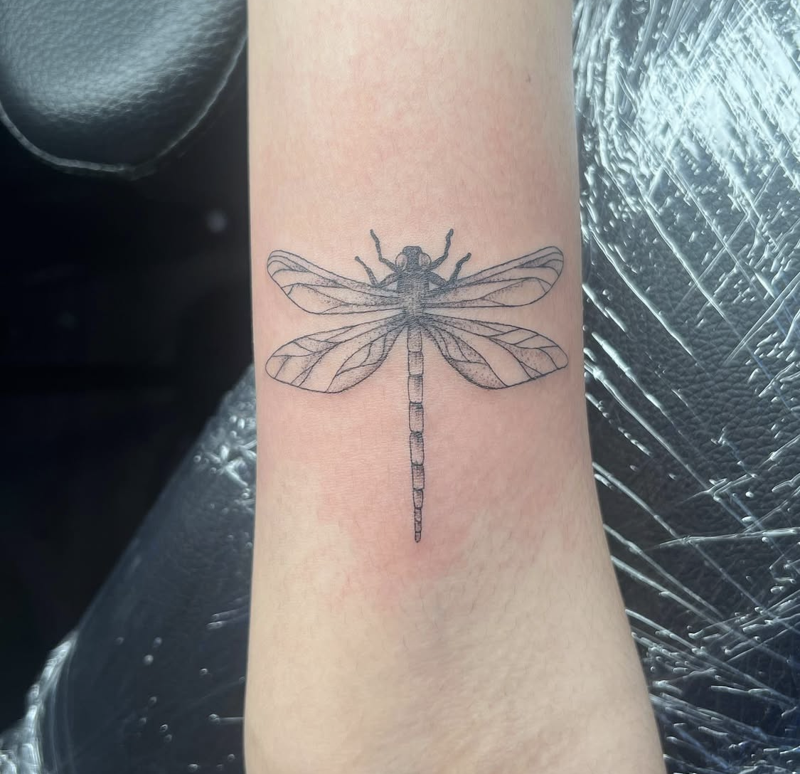 Tattoo of a dragonfly on a person's forearm, showing detailed line work of its wings, body, and head.