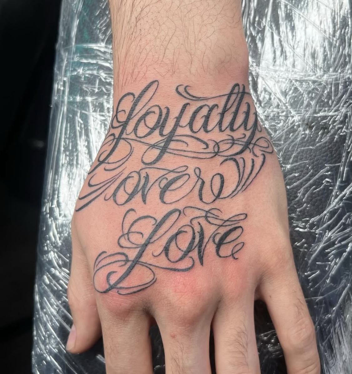 Tattoo on hand reading, "loyalty forever love" in elegant cursive script.