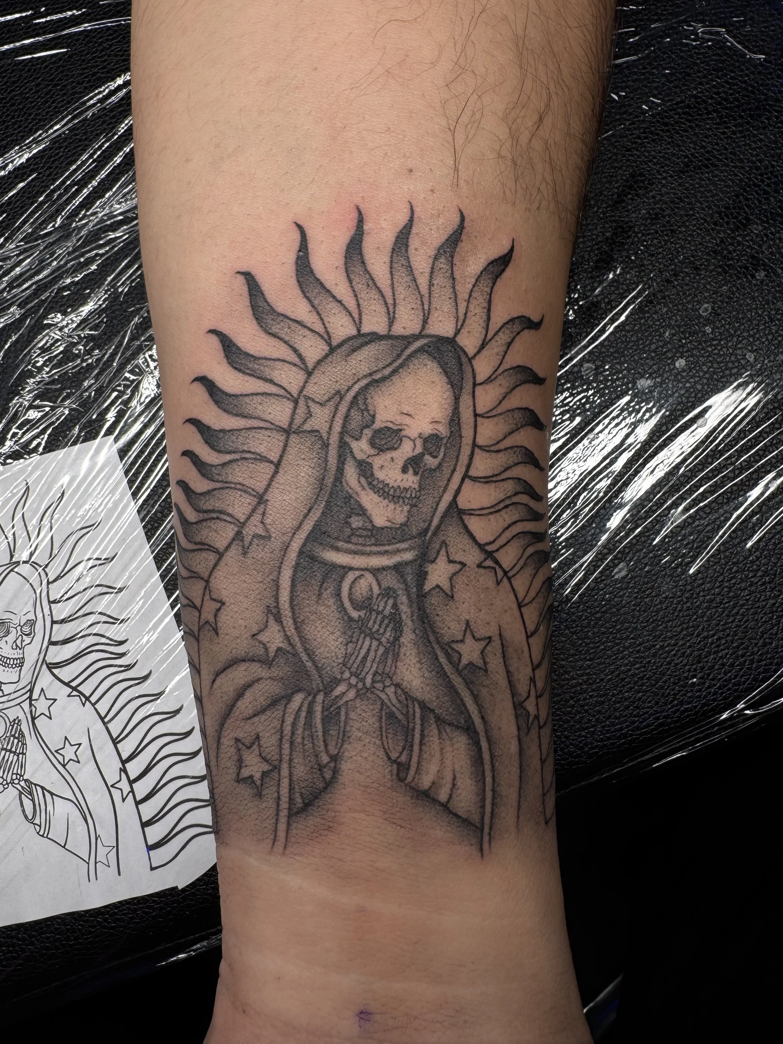 Tattoo of a skeleton face inside a religious icon with hands clasped, surrounded by stars and a halo of flames.