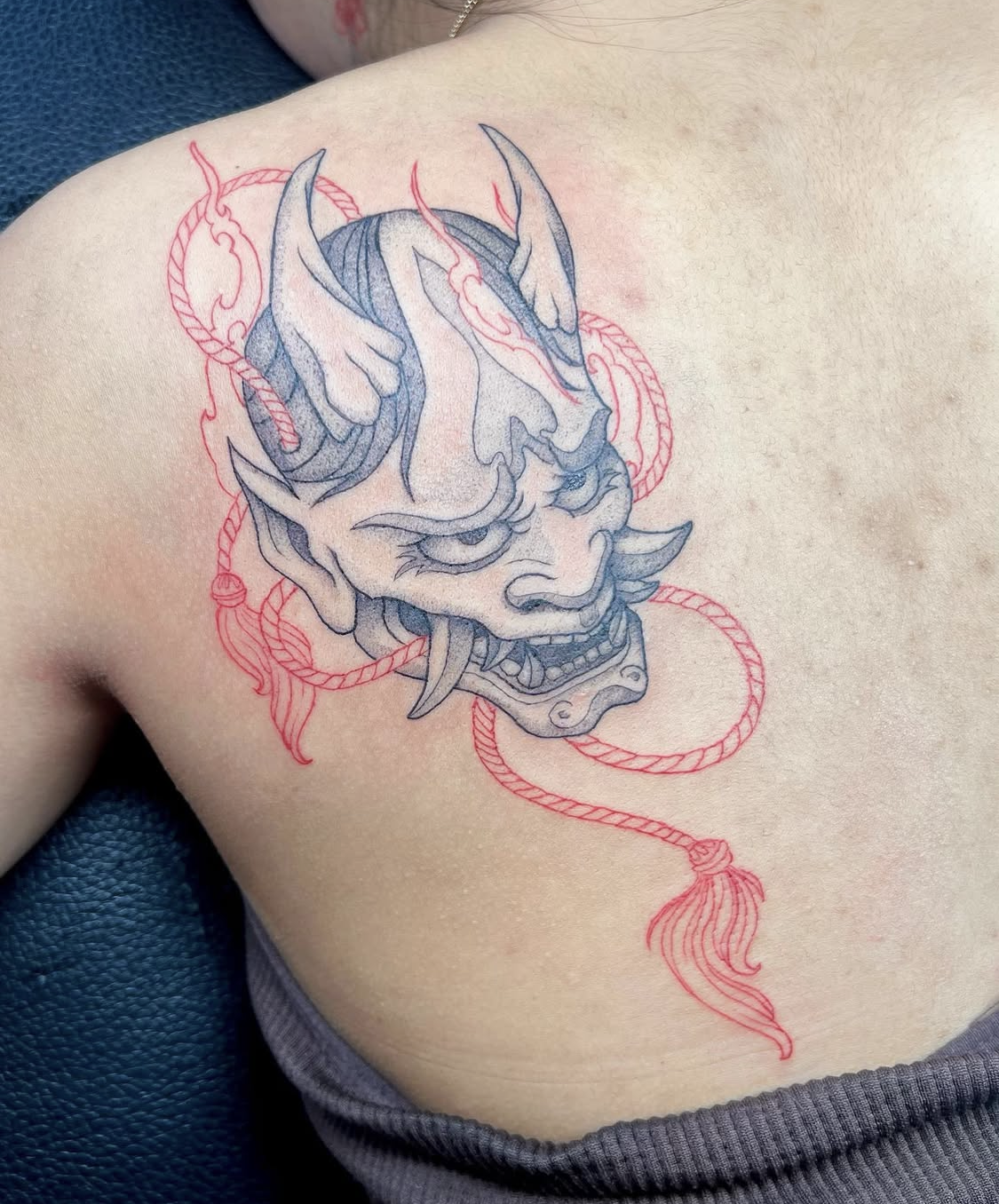 Tattoo of a blue and gray demon or oni face with horns, fierce expression, and sharp teeth, surrounded by red rope detailing on the shoulder.