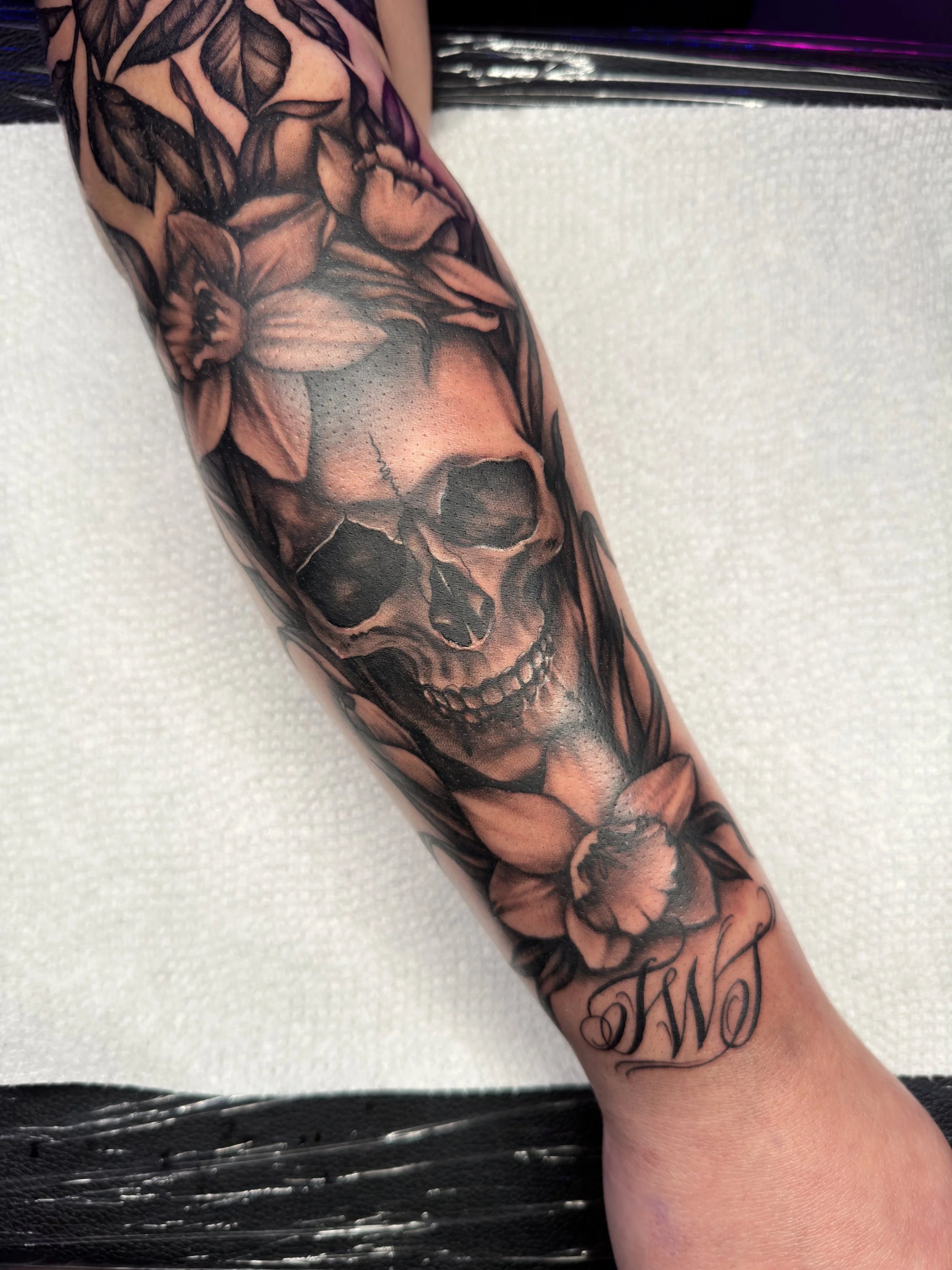 A black and gray tattoo sleeve featuring a skull surrounded by flowers and leaves, with a script that says "TWO" beneath it.