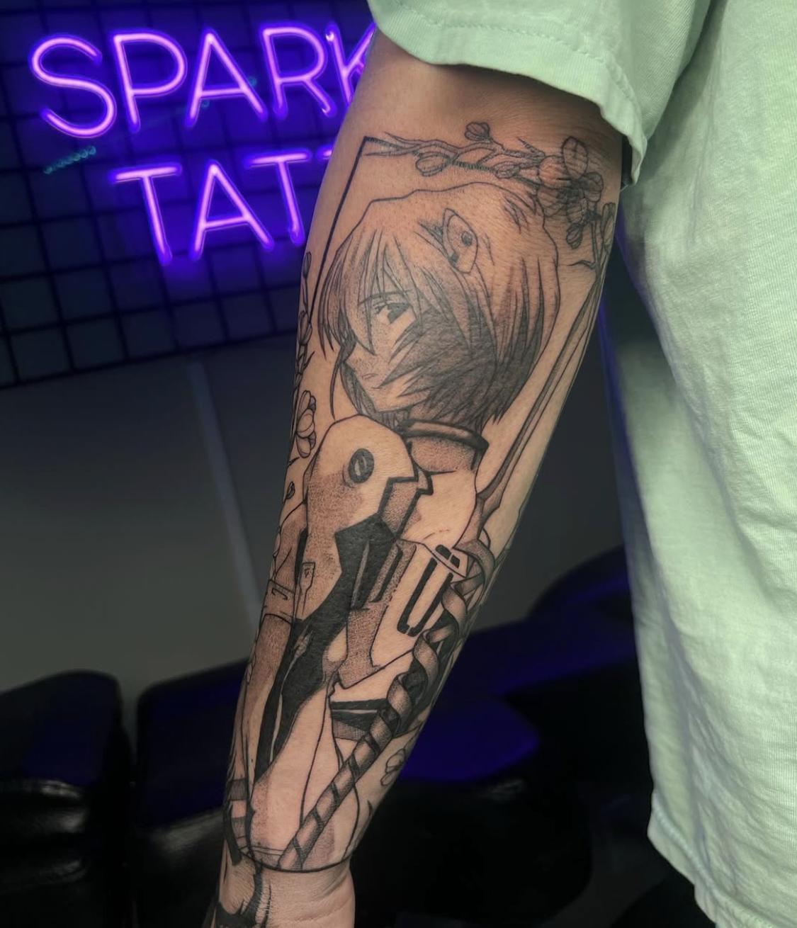 Black outline tattoo on forearm of a girl with short hair and elf ear, surrounded by flowers and a plant, with a neon purple sign in the background reading 'SPARK TAT'.