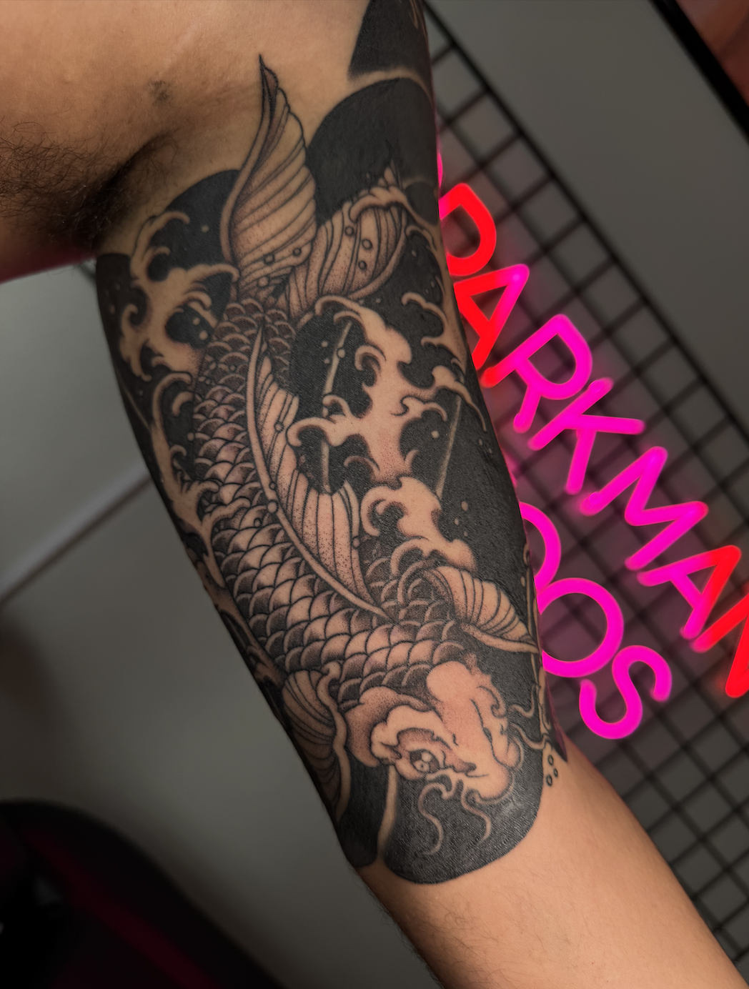 Black and gray tattoo of a koi fish swimming among waves and clouds on a person's arm, with the background showing a pink neon sign that reads 'DARKMAN LAS VEGAS'.