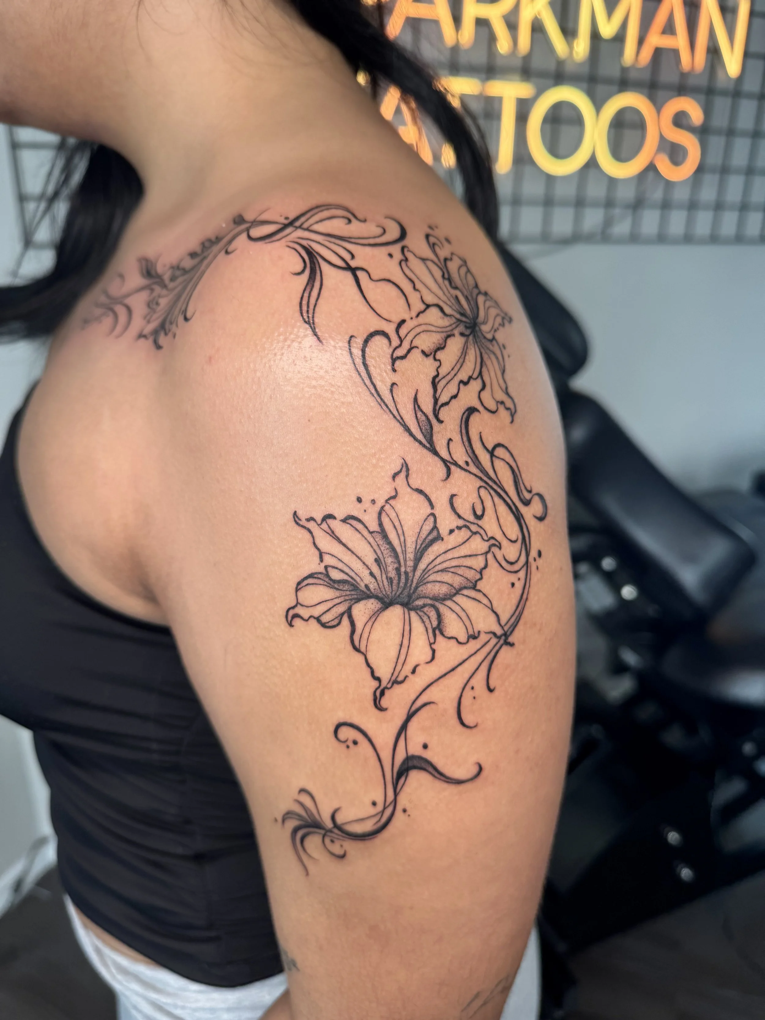 Black line floral tattoo on shoulder and upper arm featuring large flowers and swirling decorative elements.