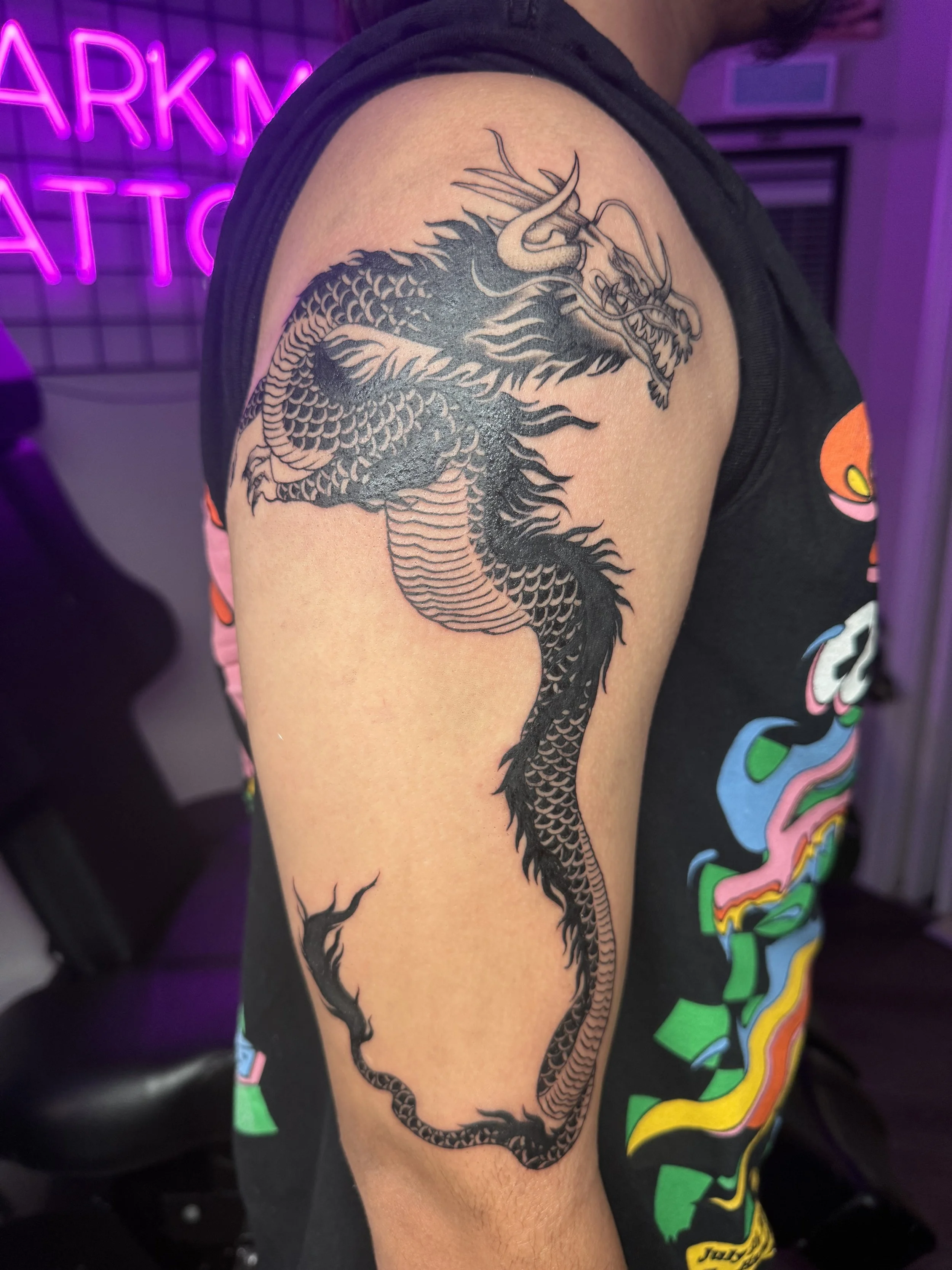 Black ink tattoo of a Chinese dragon on person's upper arm, featuring detailed scales, horns, and a fierce expression, with flames and cloud motifs.