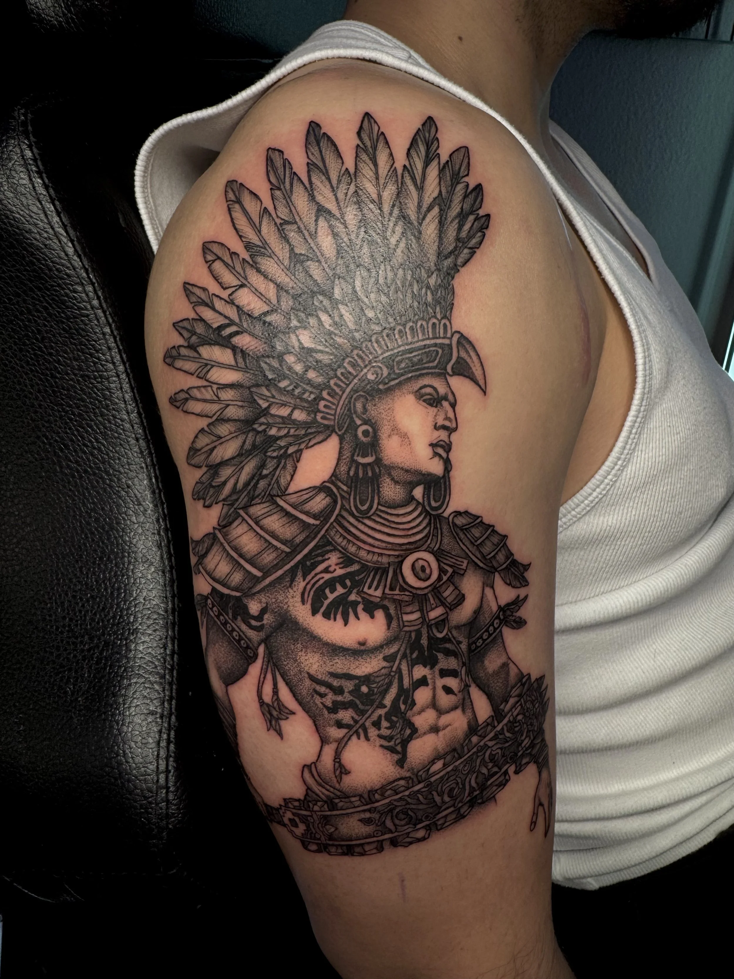 Black and gray tattoo of an ancient warrior in a headdress with feathers, armor, and necklaces on a person's upper arm.