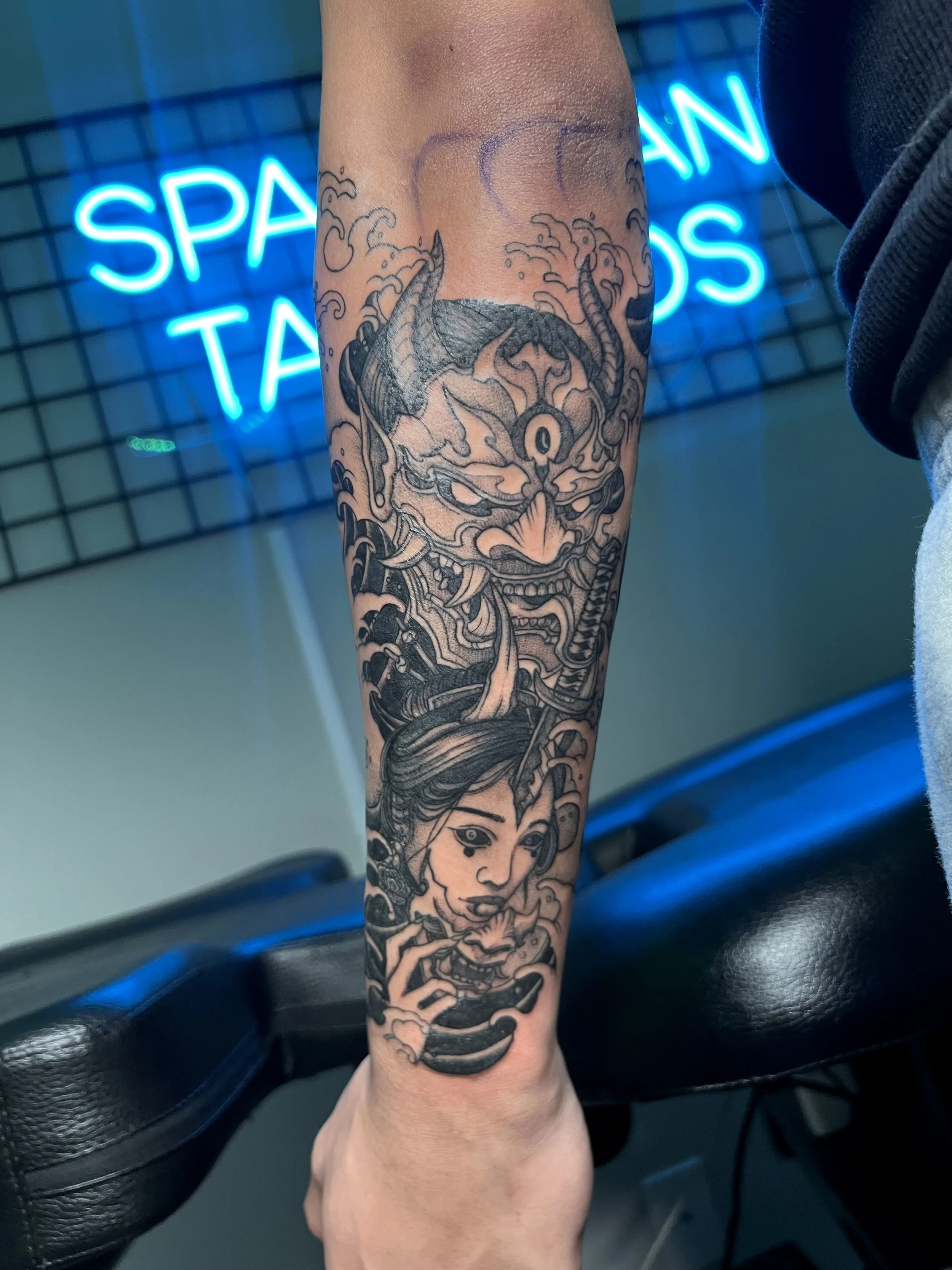 Black and gray tattoo of a fierce demon face above a serene woman with a unicorn horn on her forehead, on a person's forearm.