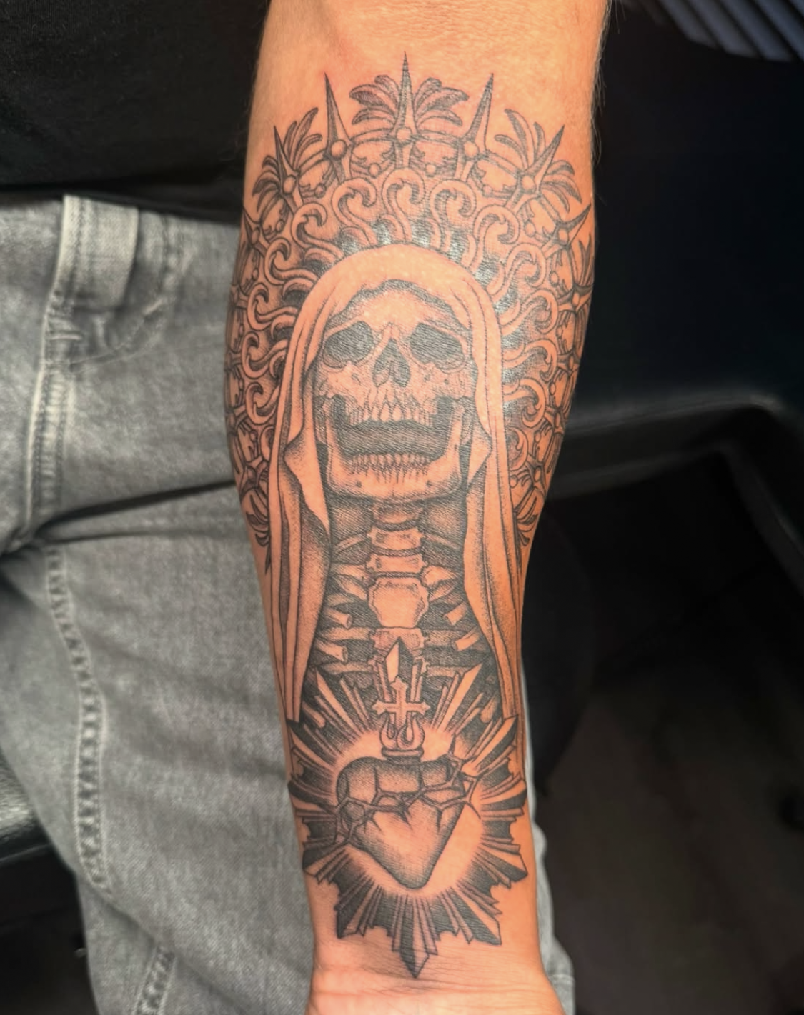 Tattoo of a skull with a hood, surrounded by a radiant halo, with a heart and dagger at the bottom.
