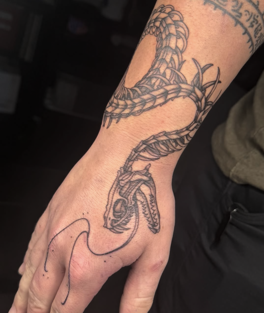 A detailed black and gray tattoo of a snake coiled around the wrist and arm, with the snake's head near the hand, featuring scales and open mouth showing teeth.
