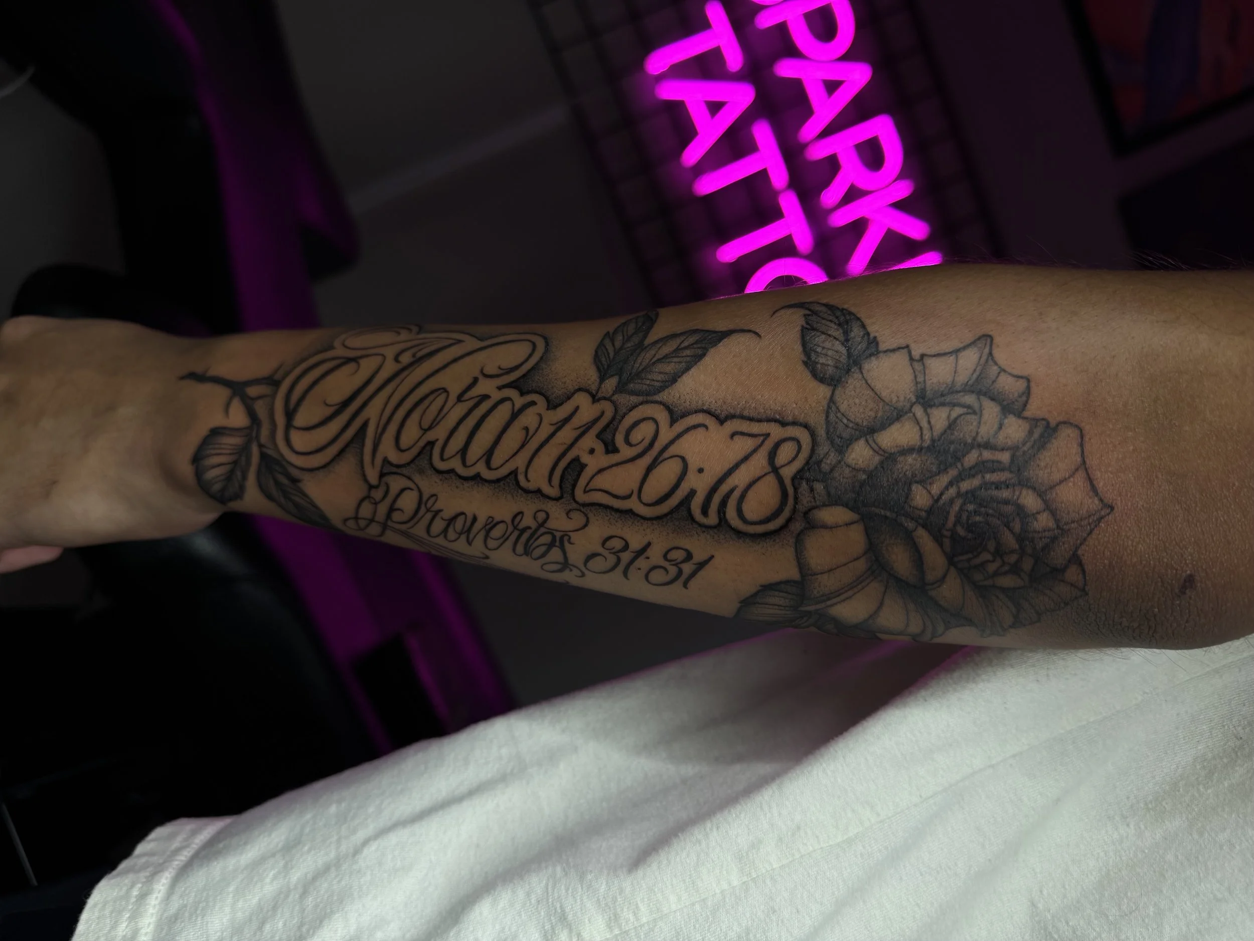 Tattoo on person's arm with the date 'January 26, 2018' and the phrase 'Proverbs 31:31,' alongside a detailed rose and leaves, with a neon pink sign with partially visible words in the background.