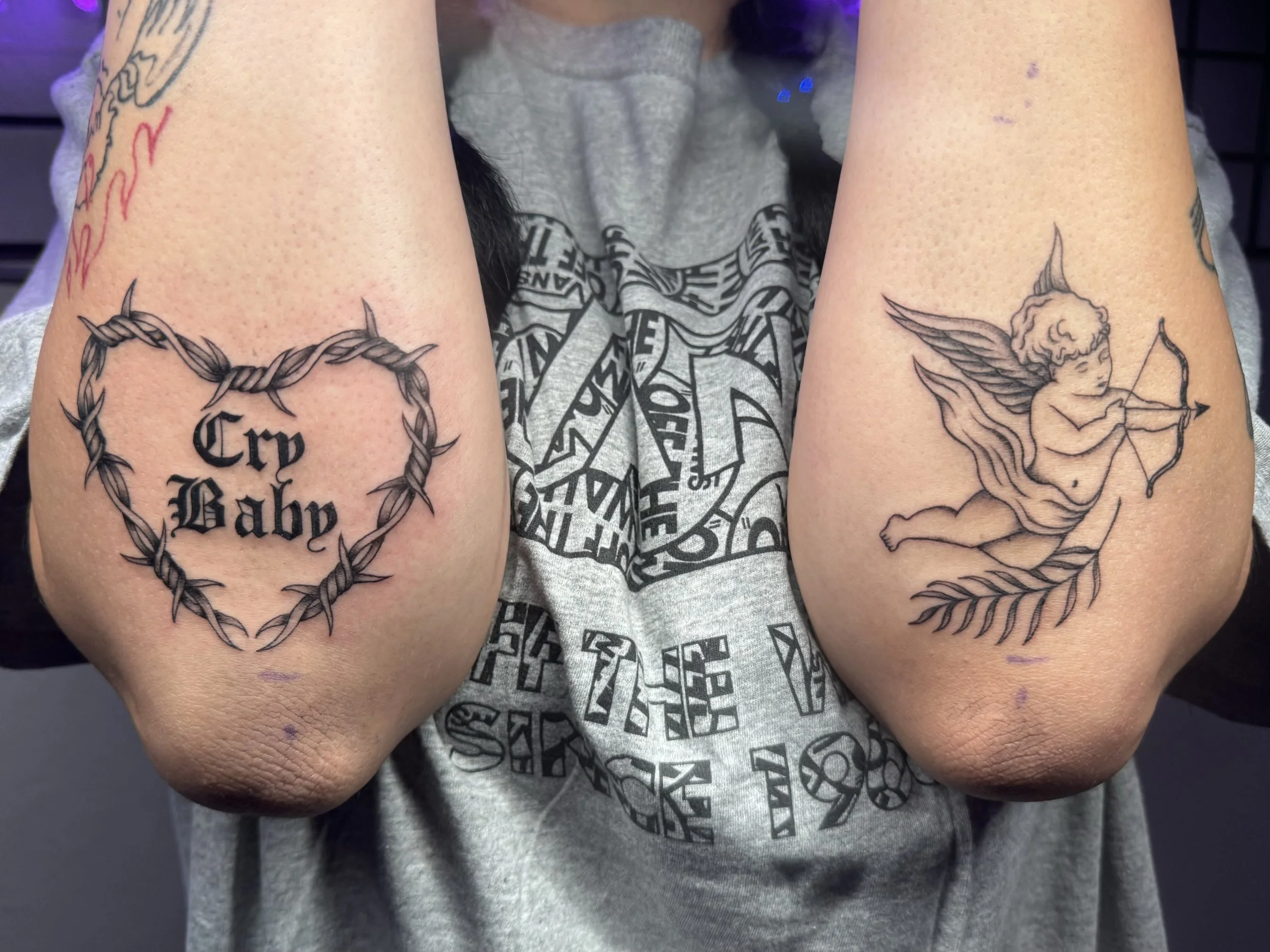 Tattoo on the person's left arm features a heart-shaped crown of thorns with the words 'Cry Baby' inside. The tattoo on the person's right arm is of a cherub with wings, holding a bow and arrow, flying over a leafy branch.