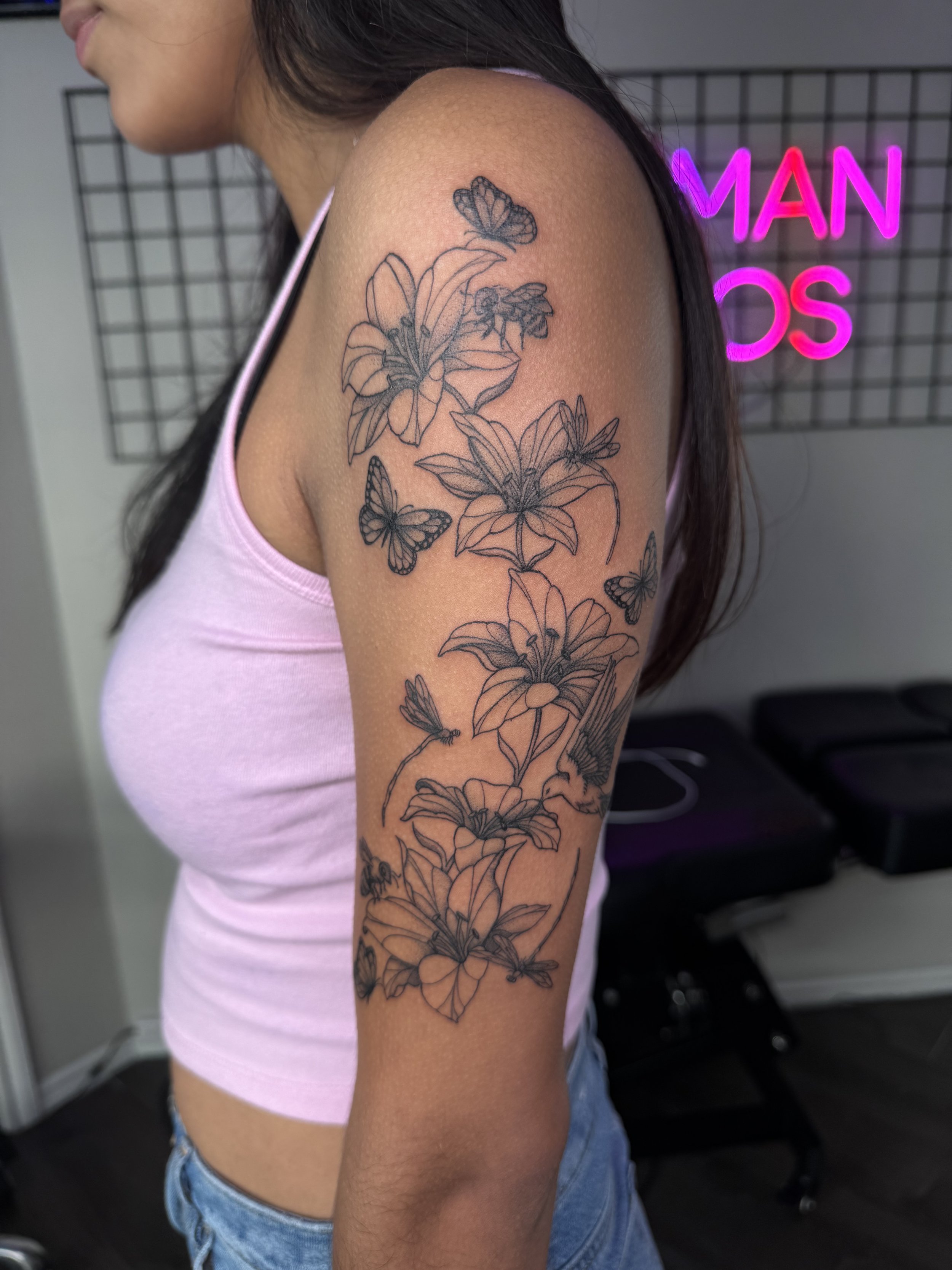 A person's upper arm with a black ink floral tattoo featuring lilies and butterflies. The person is wearing a light pink tank top and has long dark hair.